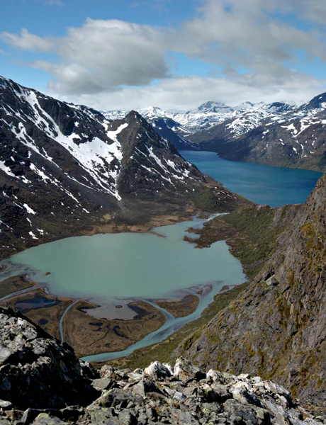Wide landscape view featuring a turquoise lake between rocky mountain slopes with patches of snow under a partly cloudy sky