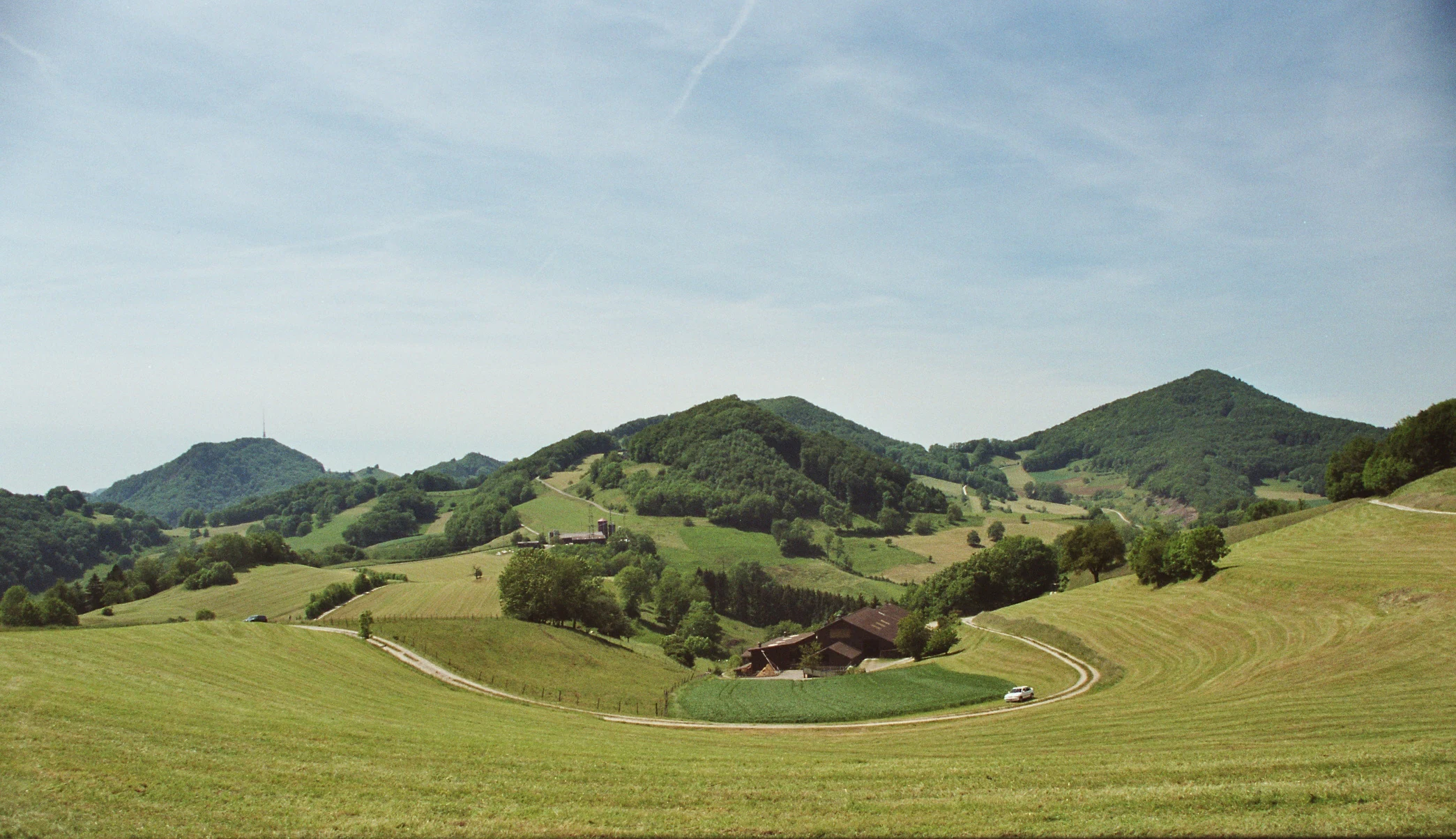 A wide landscape view of rolling green hills, forests, and grassy meadows with a winding road and a small building in the distance under a blue sky.