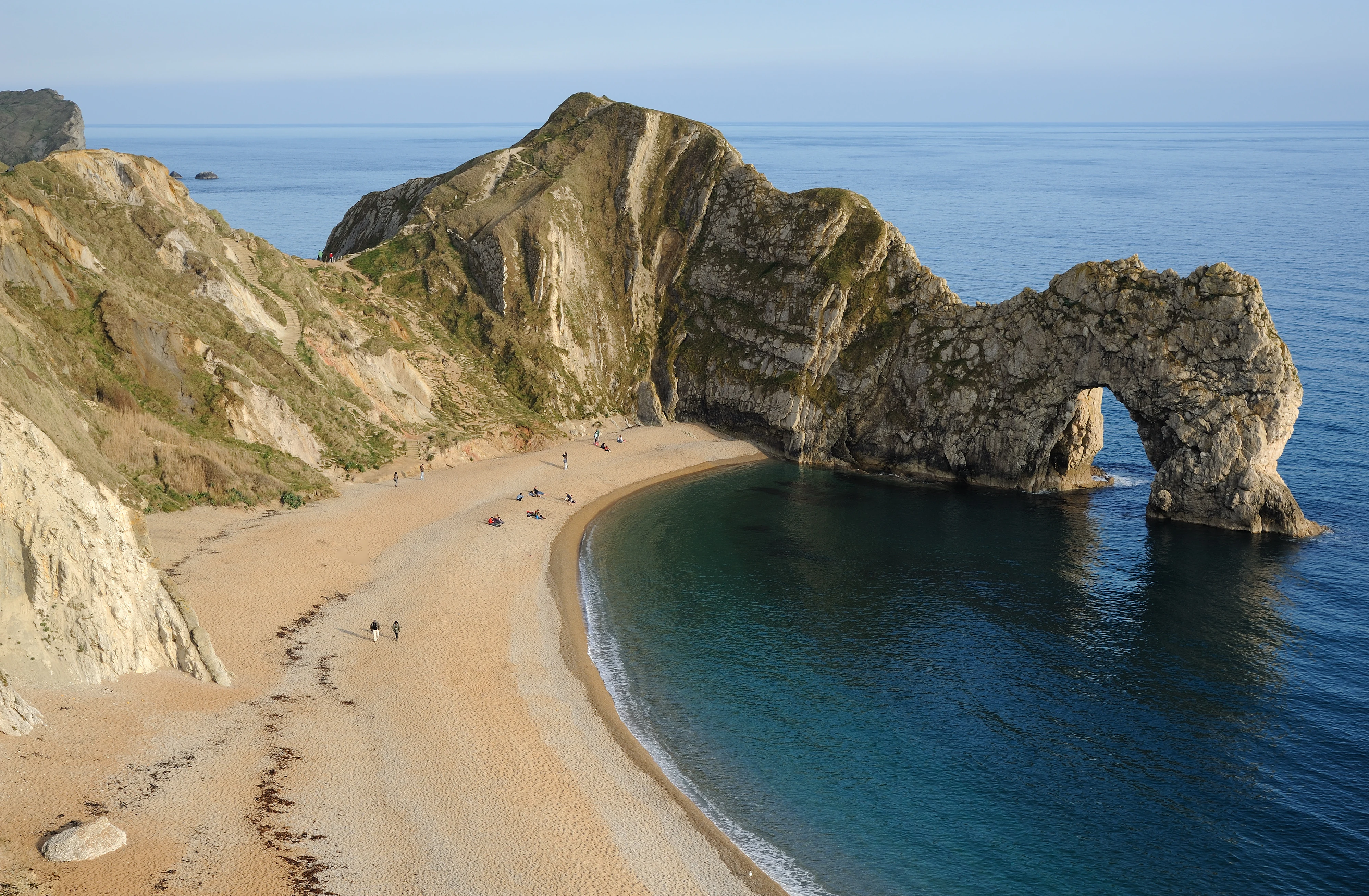 High angle shot of the natural limestone arch known as Durdle Door standing in the blue water off a curved pebble and sand beach.