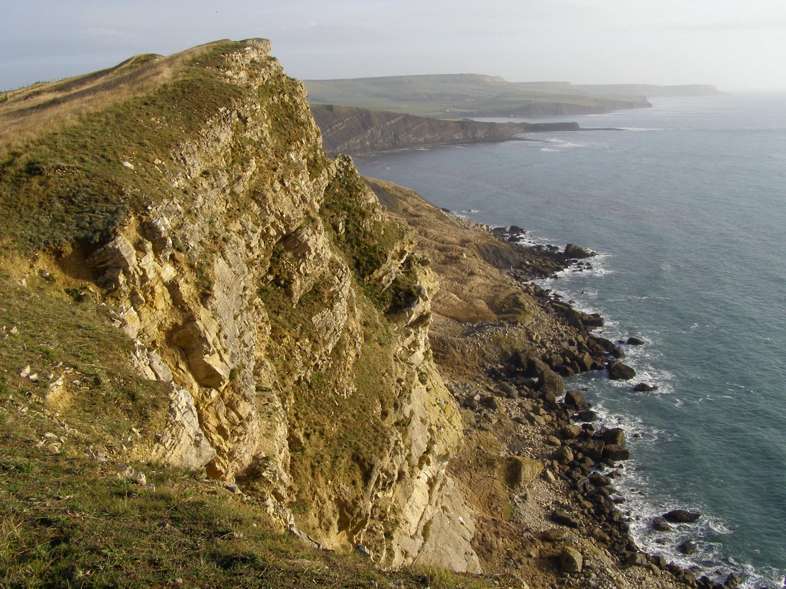 A high-angle view looking down at a steep, golden-hued rocky coastal cliff that drops sharply into the dark blue English Channel, with a distant headland stretching out into the water.