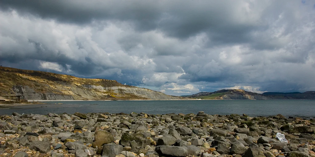 A wide view of a bay with a rocky foreground shoreline and distant coastal cliffs under a cloudy, dramatic sky.