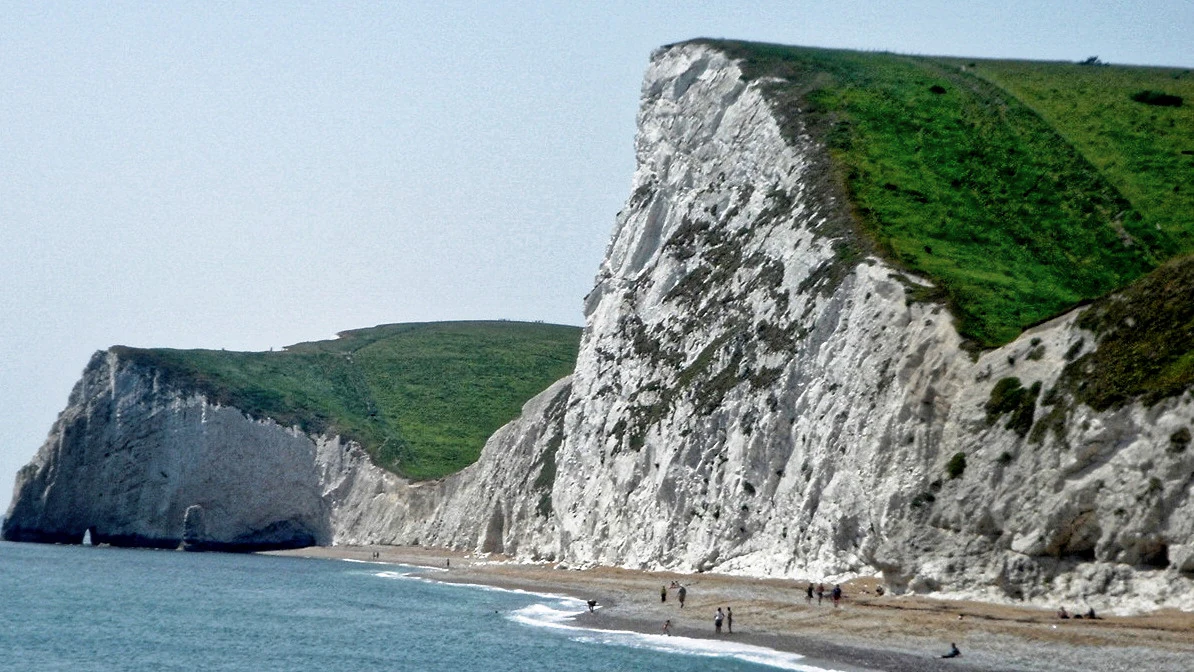 A wide angle view of white chalk cliffs towering over a pebble beach and the sea on a clear day.