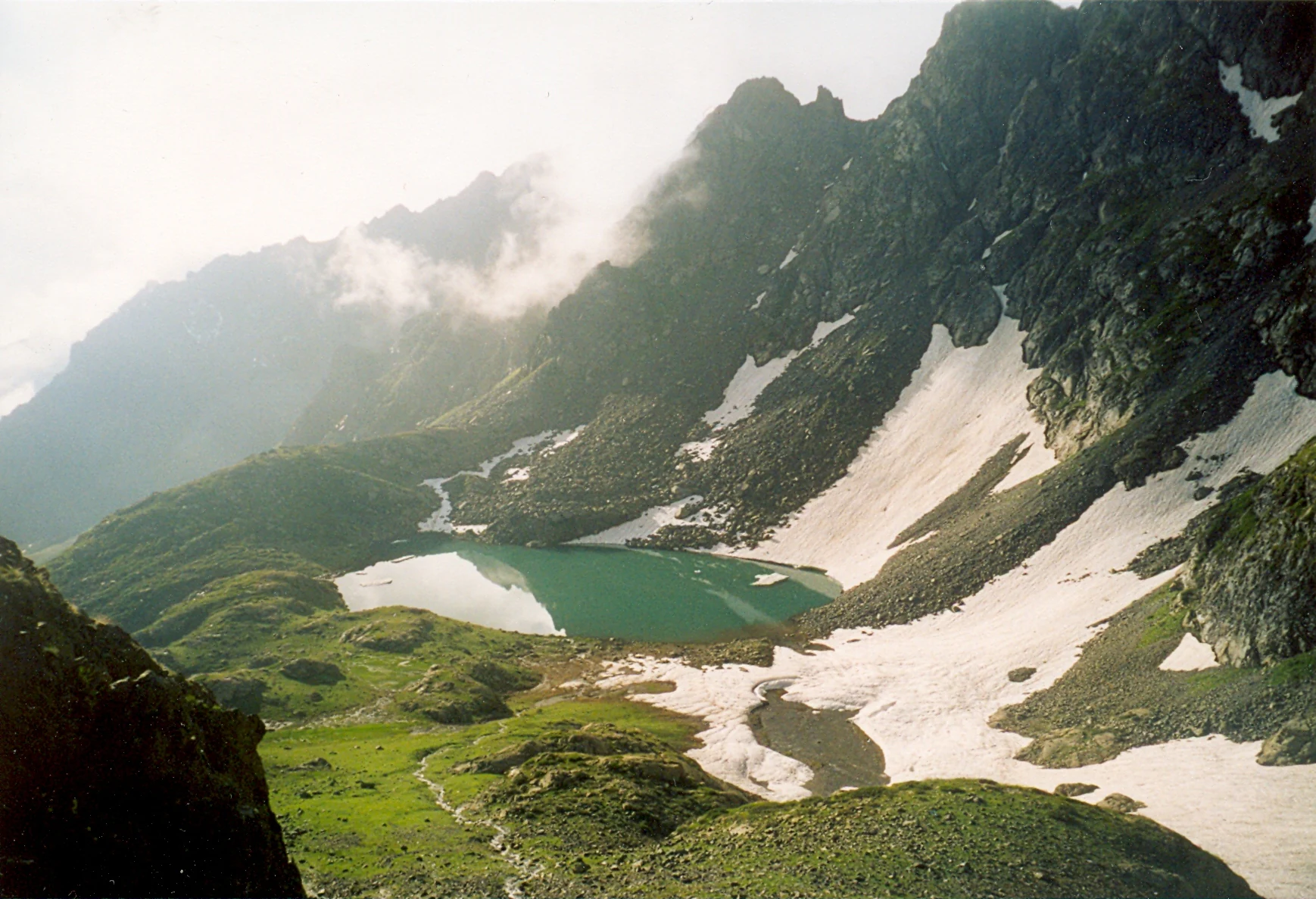 A mountain lake with turquoise water surrounded by rocky peaks with patches of snow, green meadows, and misty sky