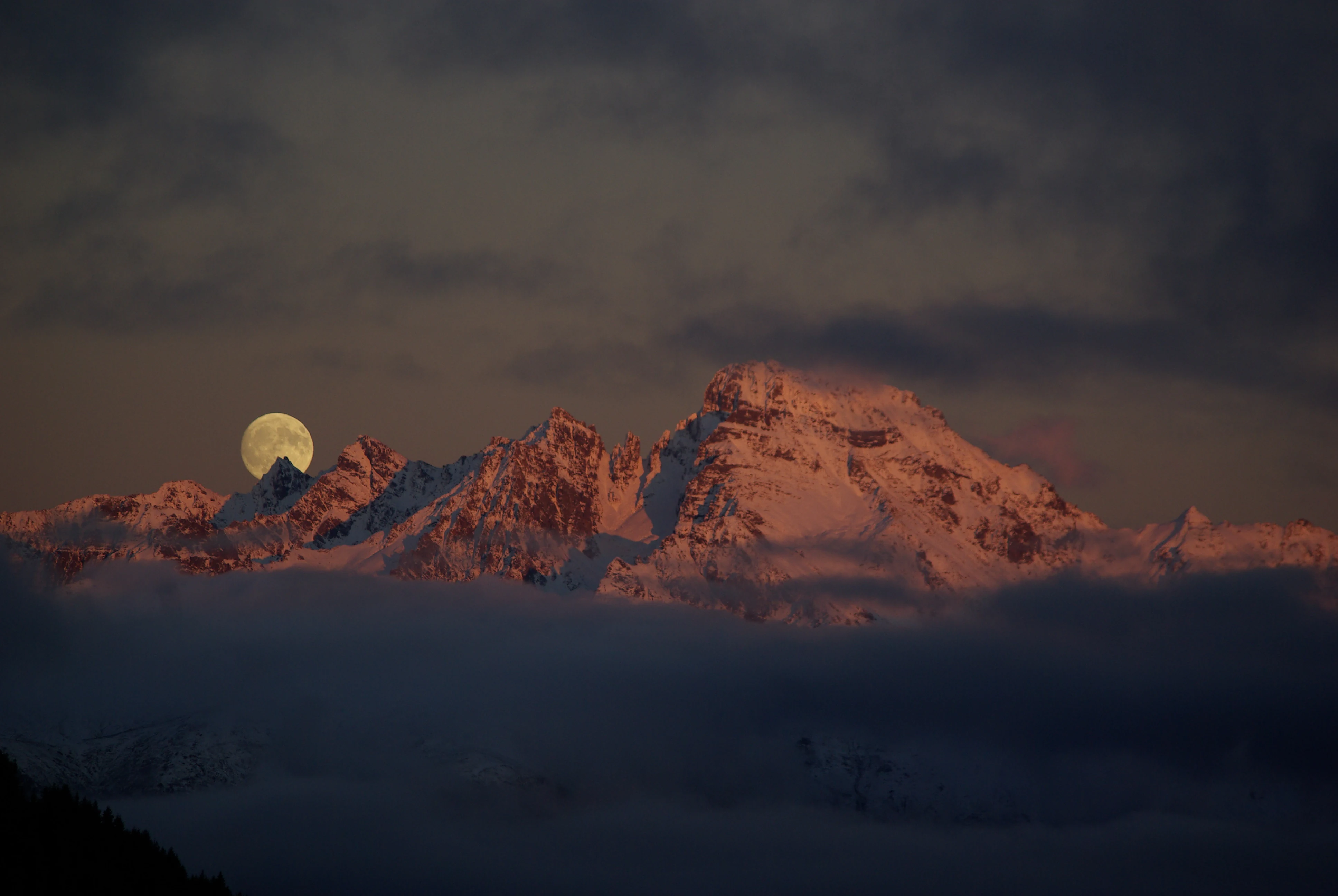 Snow-covered mountain peaks under twilight sky with full moon visible above mountains and misty clouds below