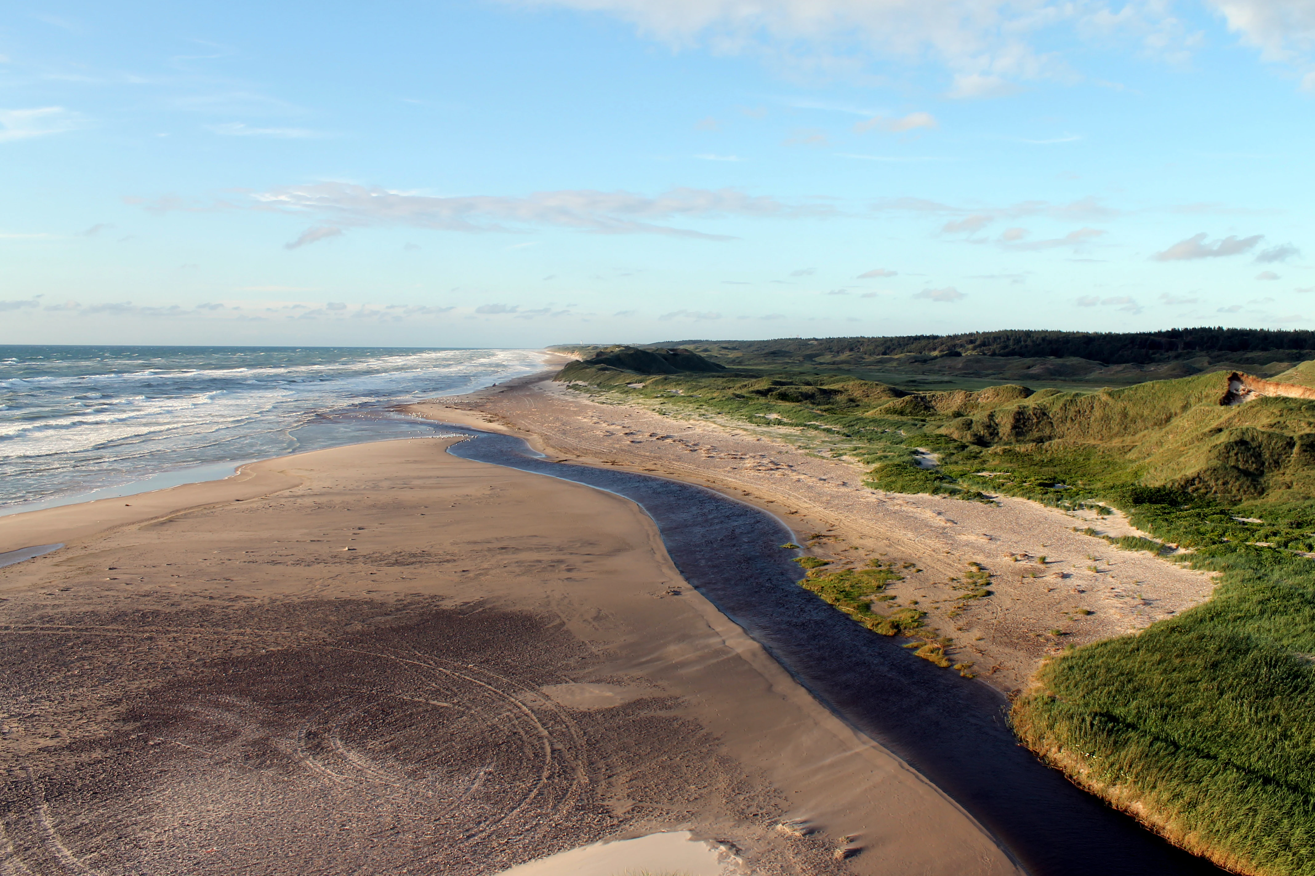 An aerial-style view of a river mouth emptying into the sea across a wide sandy beach, with vegetated dunes on the right.