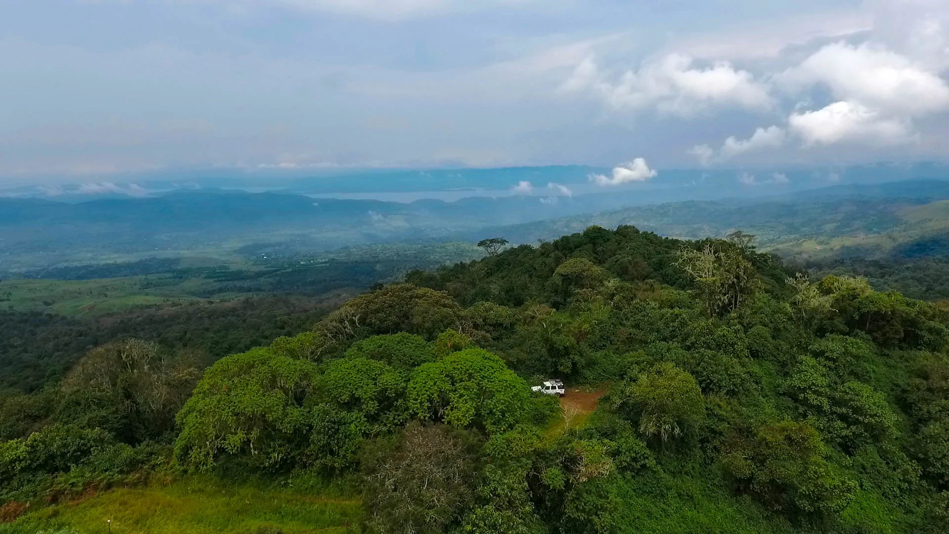 Vista aérea de un denso bosque verde con un vehículo en la cima de una colina, rodeado de colinas onduladas bajo un cielo parcialmente nublado