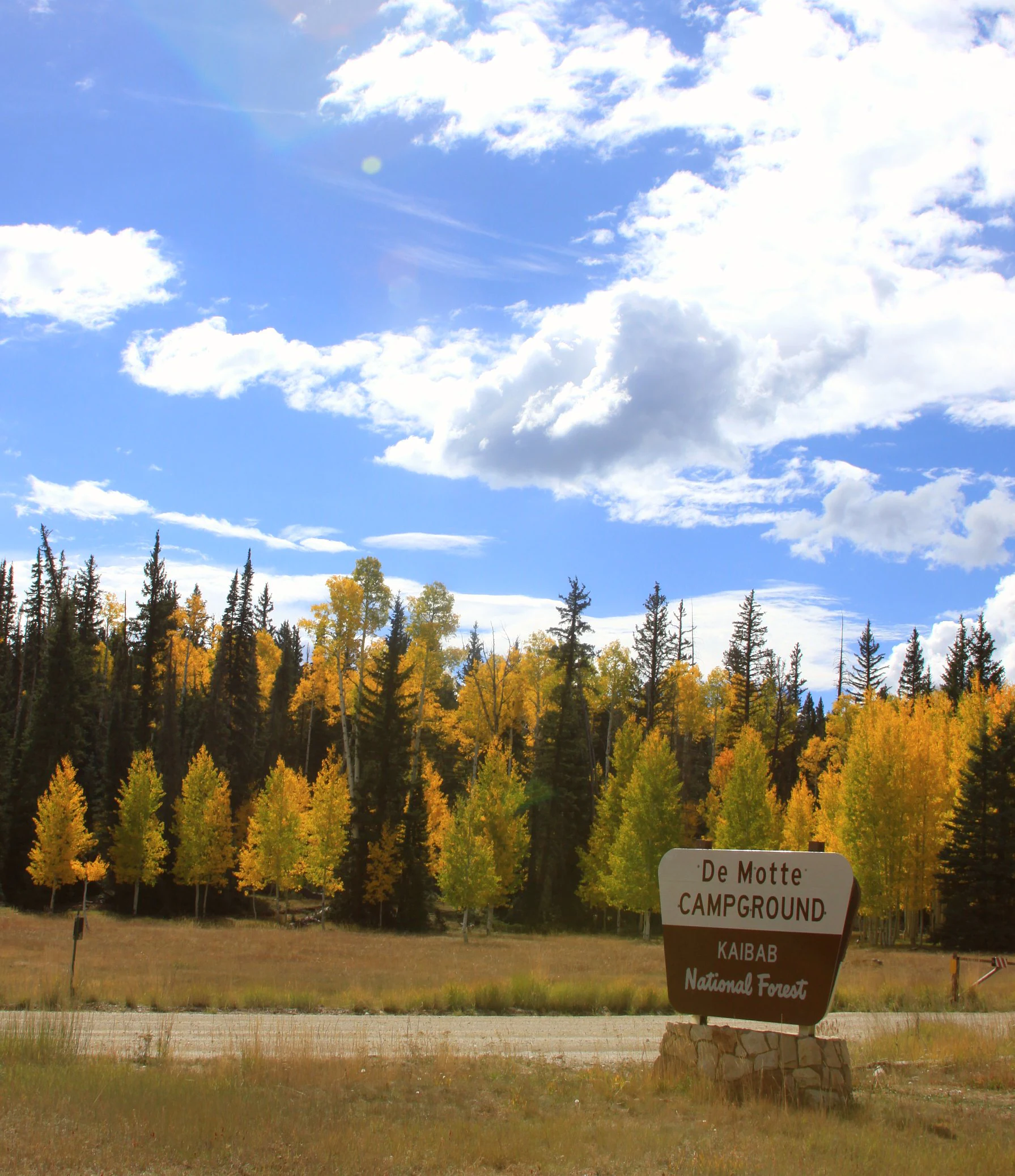 A brown and white entrance sign for De Motte Campground in Kaibab National Forest stands in the foreground, with a golden aspen and evergreen forest in the background under a blue sky with scattered clouds.