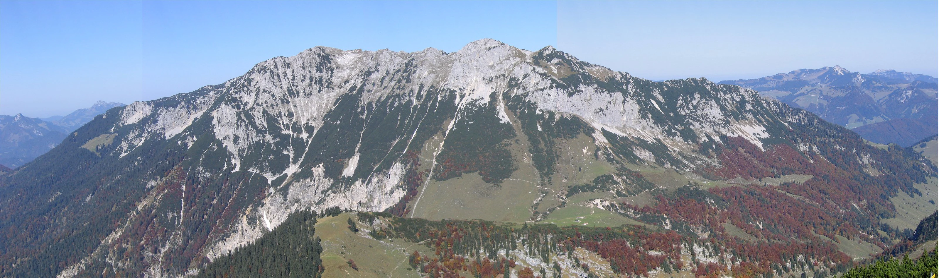 A wide, panoramic shot showing the rugged limestone peaks of the Zahmer Kaiser mountain range, characterized by rocky slopes, coniferous forests, and autumn-colored foliage.