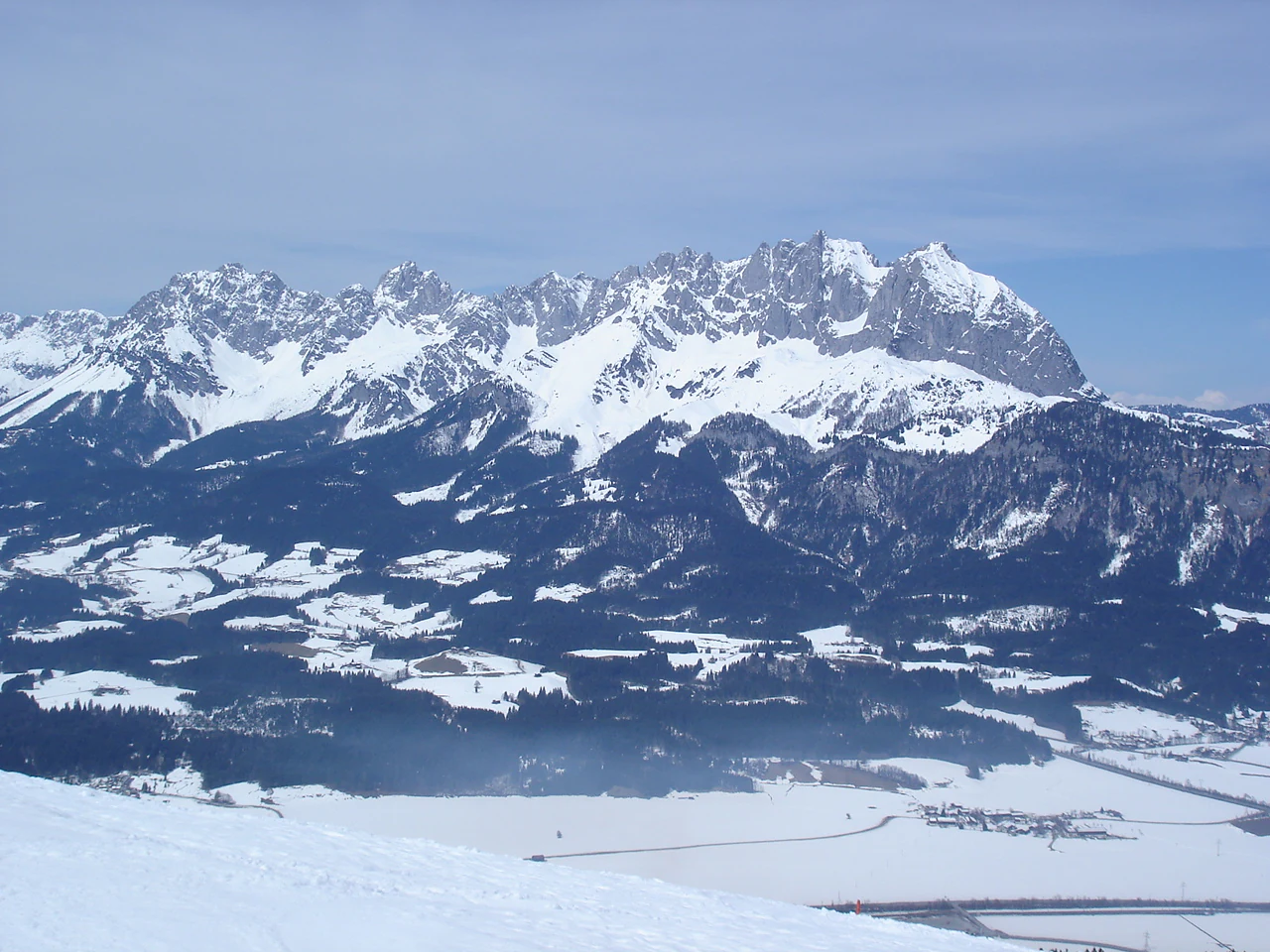 A wide, eye-level landscape shot of a rugged, snow-dusted mountain range rising above a snowy valley floor under a clear sky.
