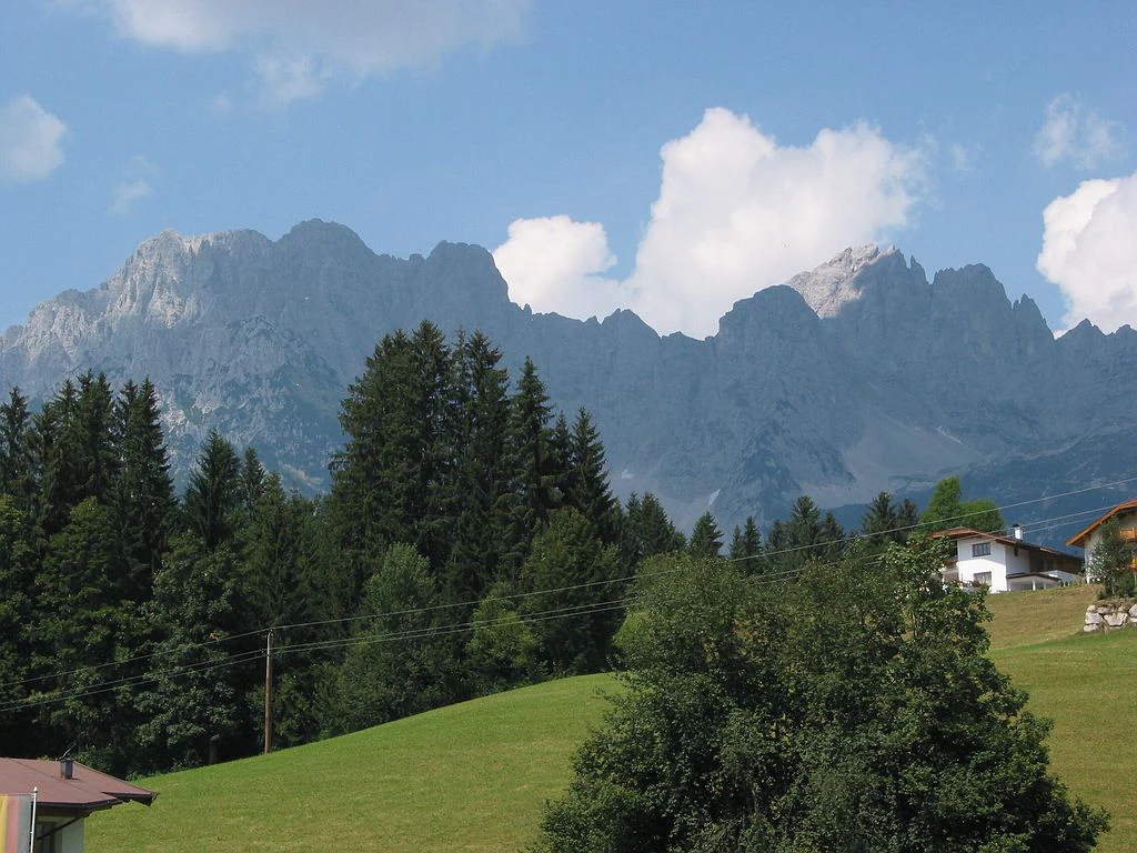 A wide scenic view of the jagged limestone peaks of the Wilder Kaiser mountains, with a green alpine meadow and coniferous forest in the foreground and a few houses on the right.