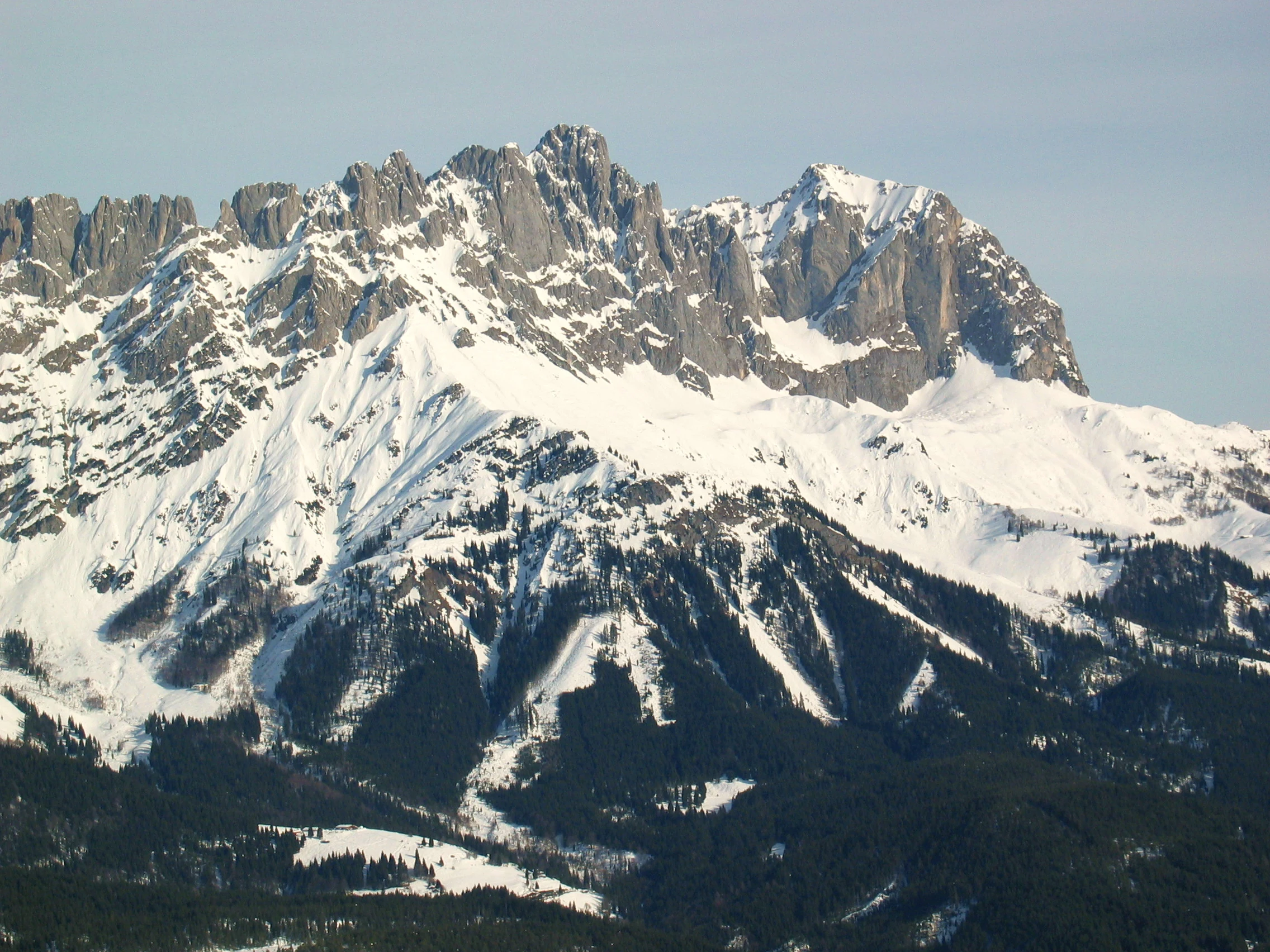 A wide, high-angle shot of rugged, snow-covered mountain peaks with steep rocky faces and dense evergreen forests at the base.