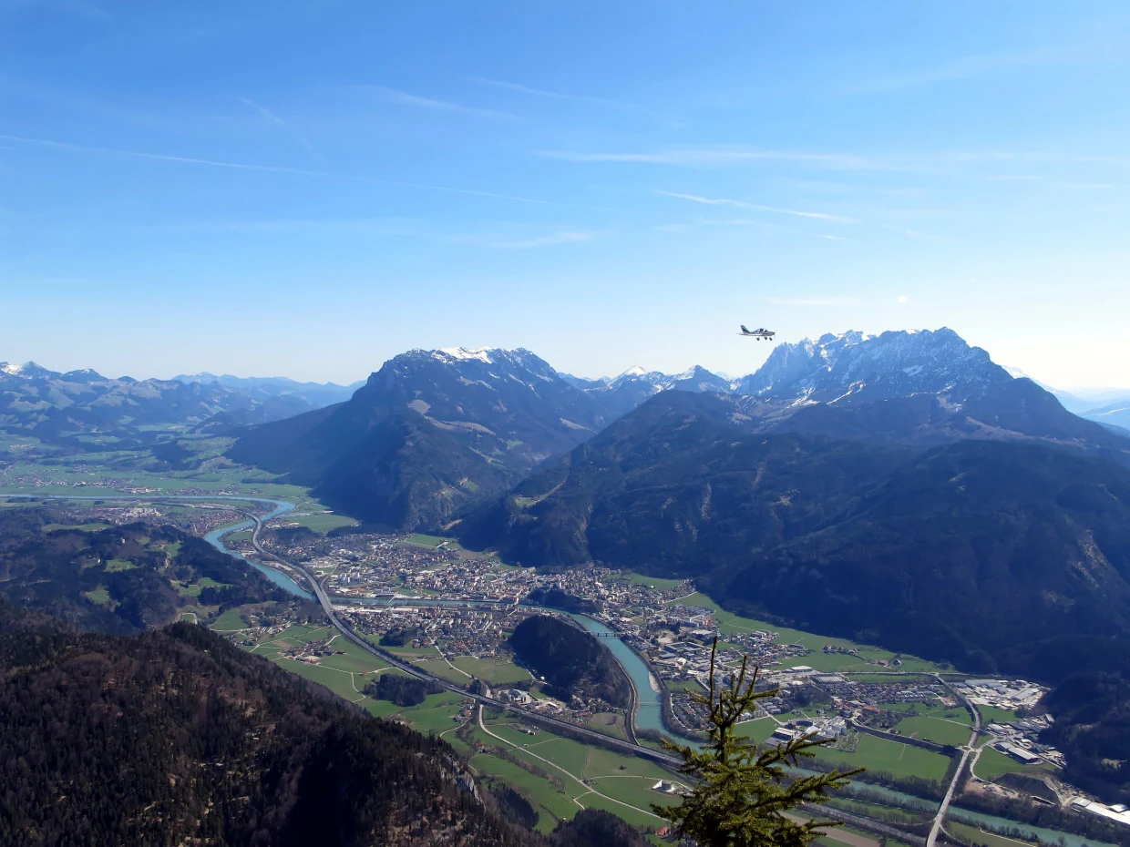 A wide, high-angle view looking down into a valley containing a town and a winding river, flanked by large, rugged mountains under a clear blue sky.