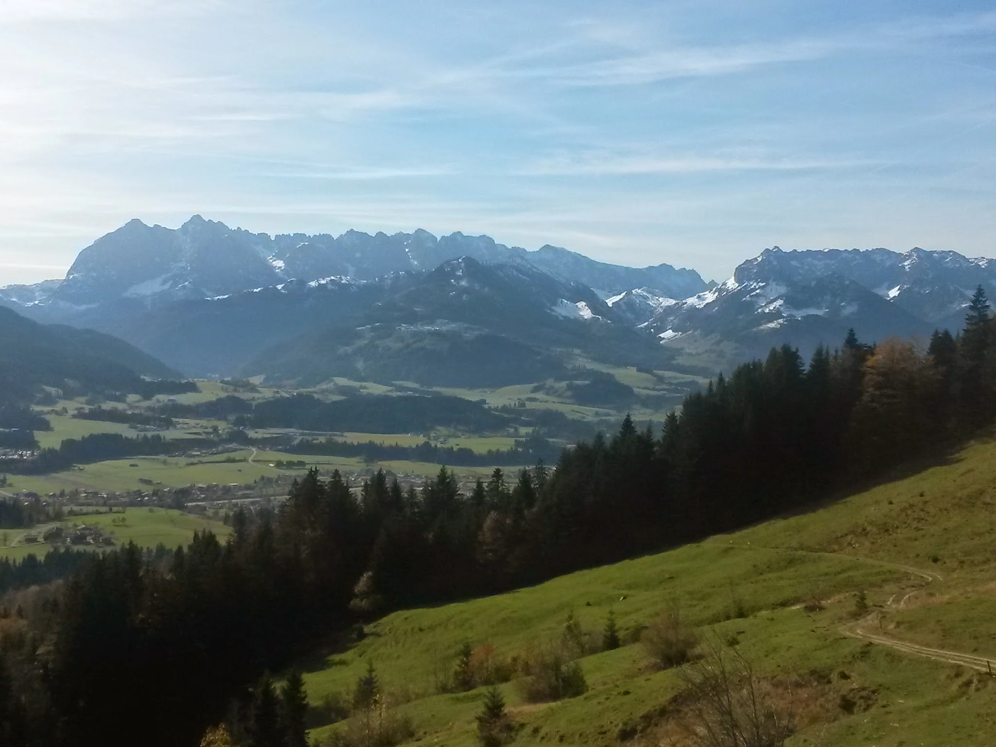 A wide landscape view of the Kaiser Mountains featuring craggy limestone peaks above a green valley and evergreen forest.