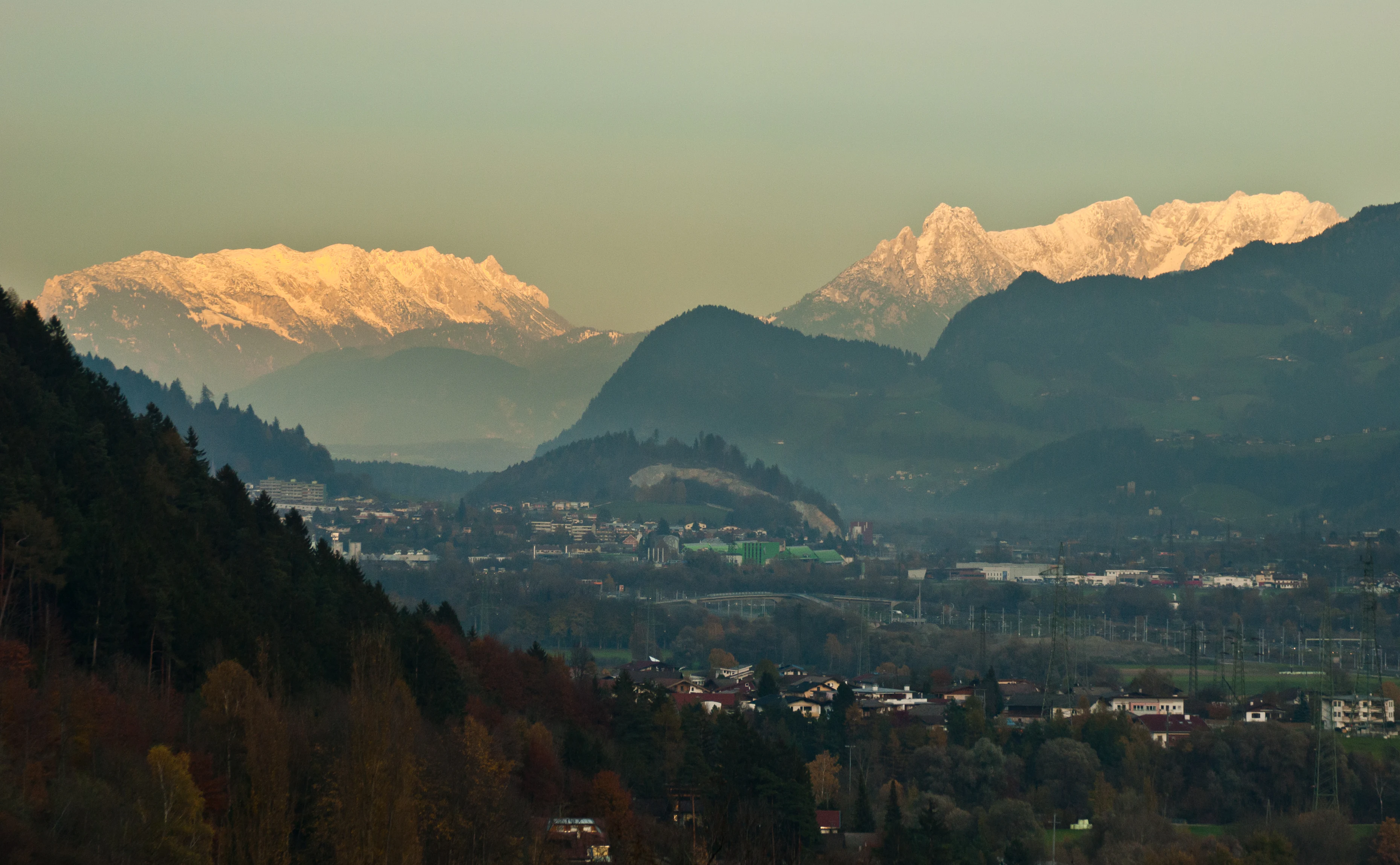 A wide mountain landscape view showing snow-capped limestone peaks of the Kaiser Mountains glowing in golden light above a valley with scattered buildings.