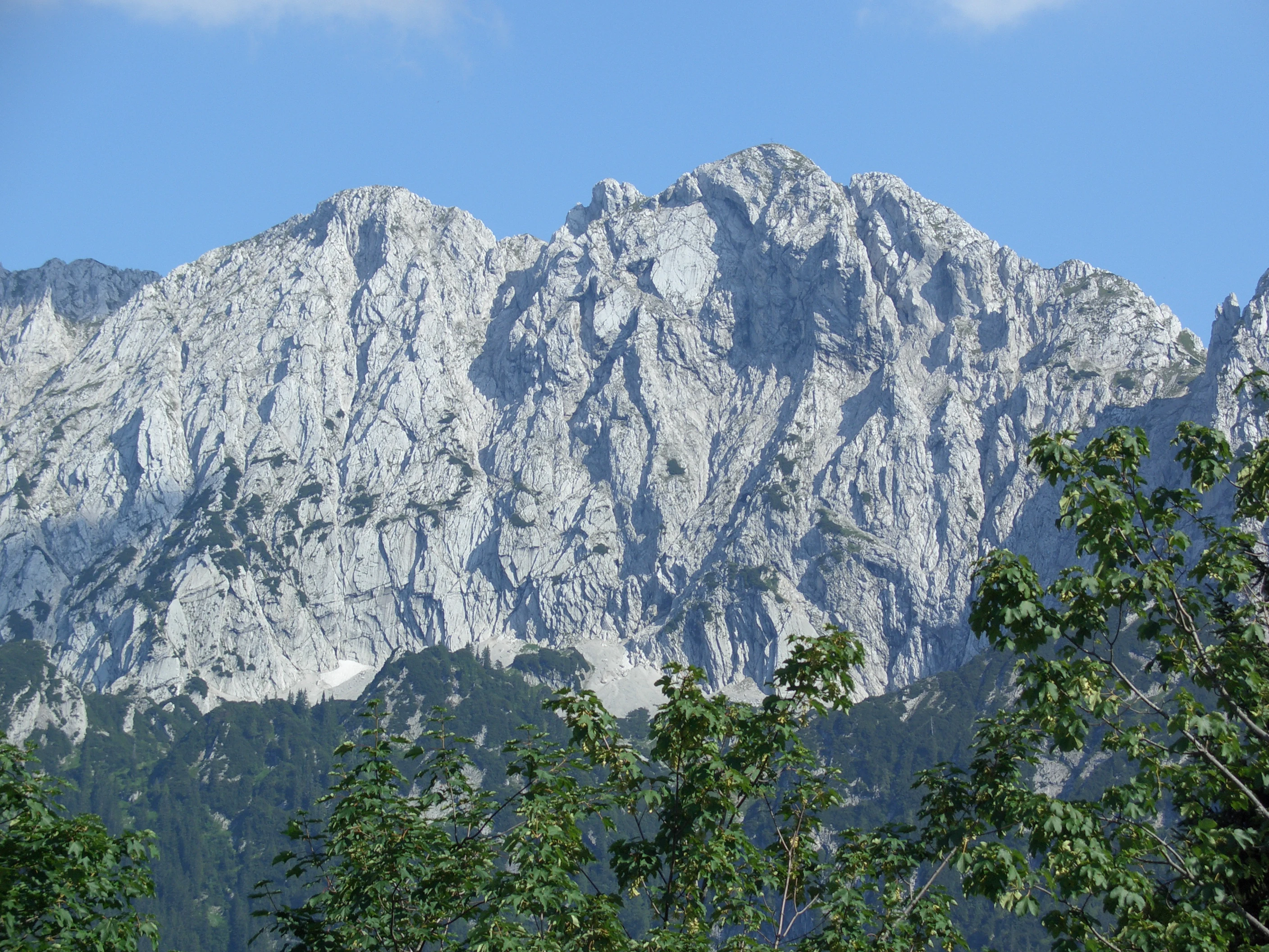 A massive limestone mountain face with rugged peaks and rocky slopes, viewed from below with green trees in the foreground against a clear blue sky.