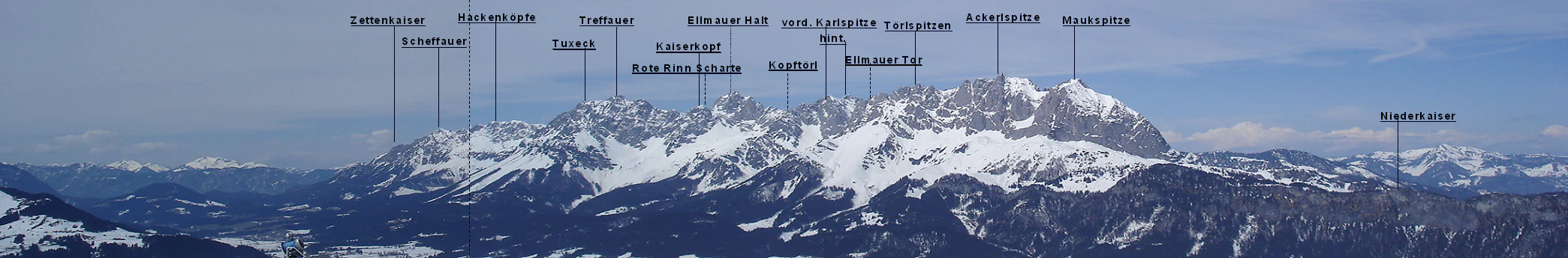A wide panoramic photograph showing a series of rugged, snow-covered mountain peaks in the Kaiser Mountains, with various summits identified by small labels and vertical indicator lines.