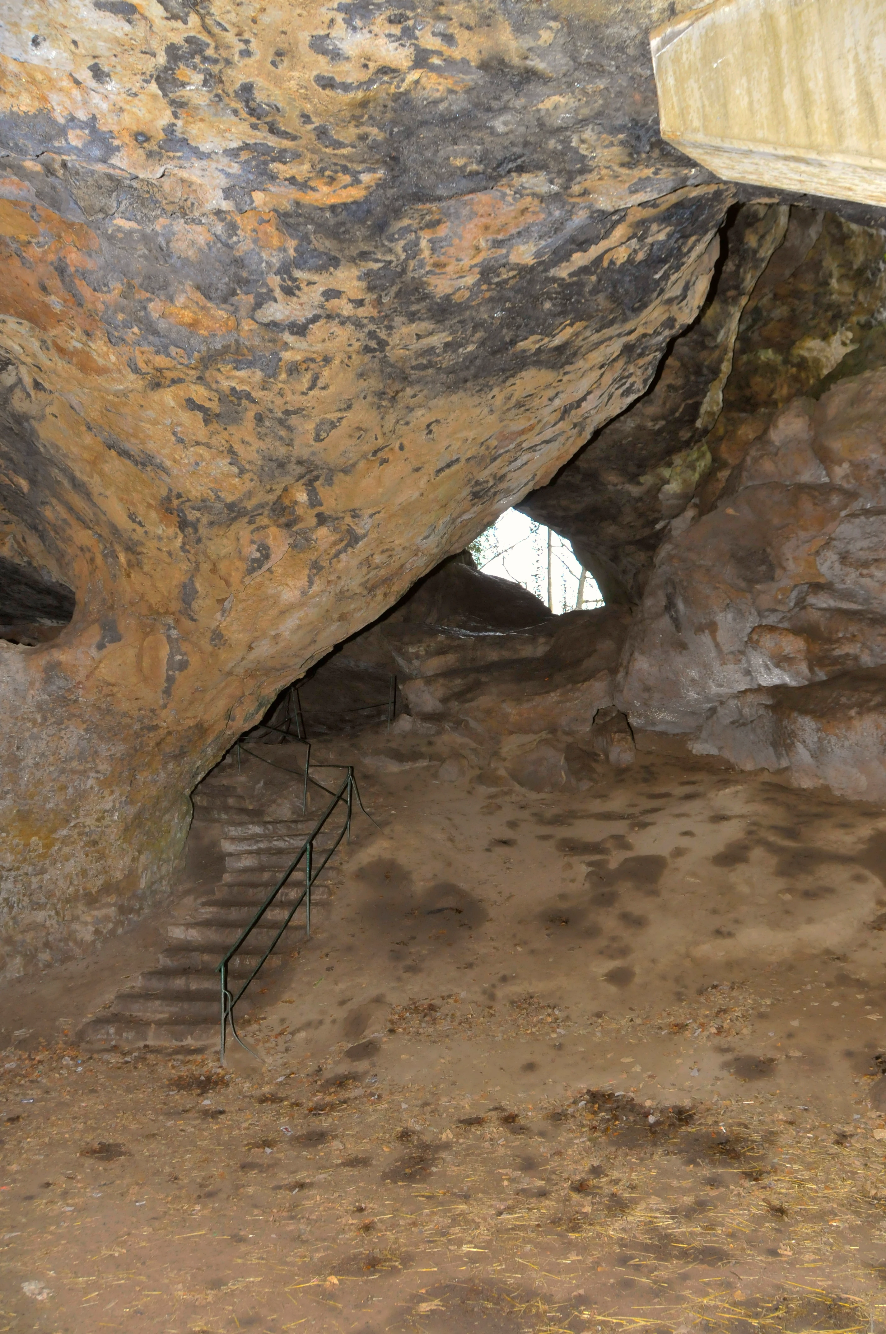 Una vista a ras de suelo dentro de la cueva Kakushöhle, que muestra una gran abertura de piedra caliza, un suelo arenoso y escalones de piedra con pasamanos metálico que conducen a lo más profundo de la caverna.
