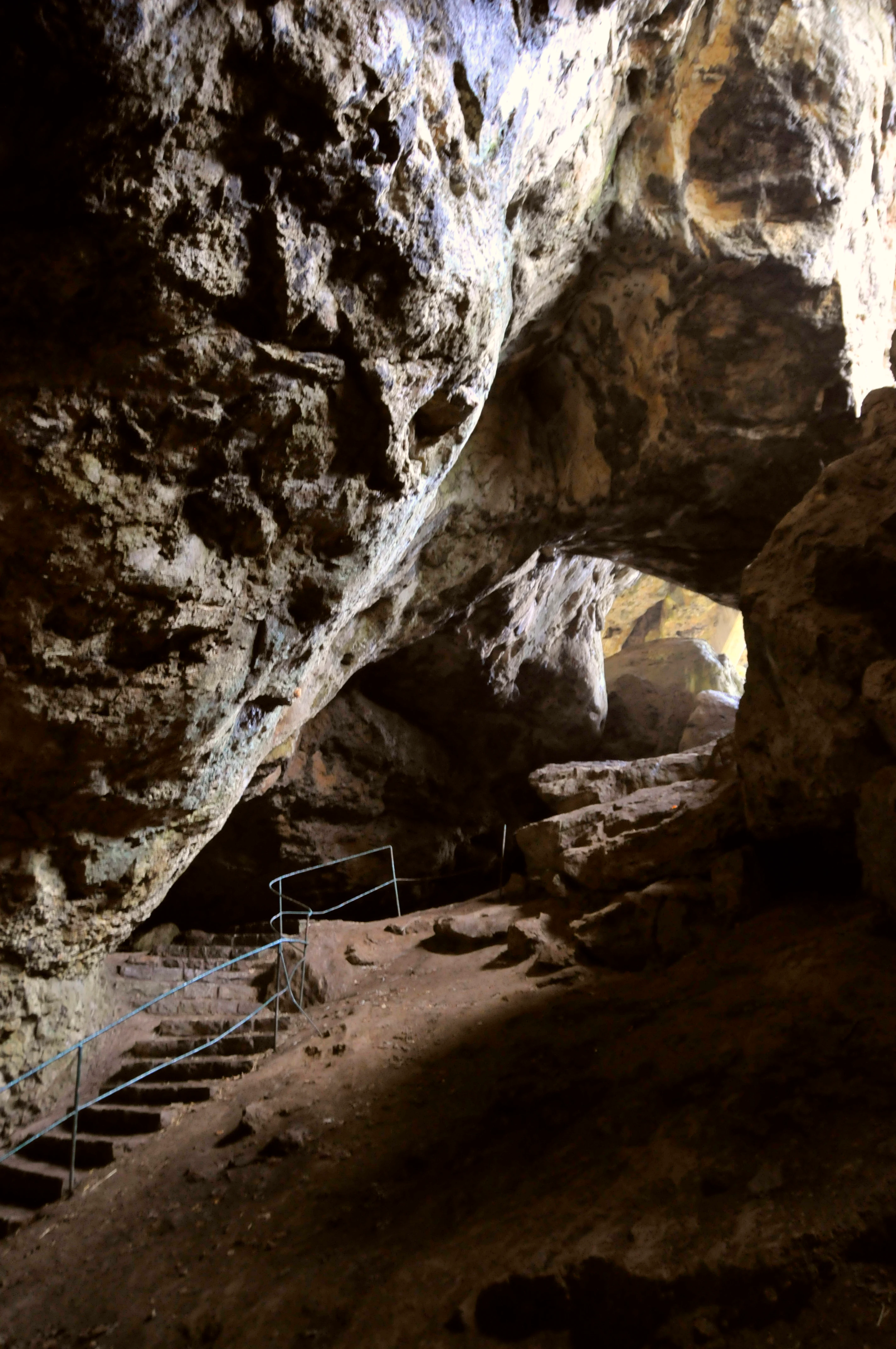 Una vista hacia la entrada de una gran cueva de piedra caliza, con paredes rocosas naturales, un sendero de tierra y un tramo de escalones de piedra con barandilla metálica.