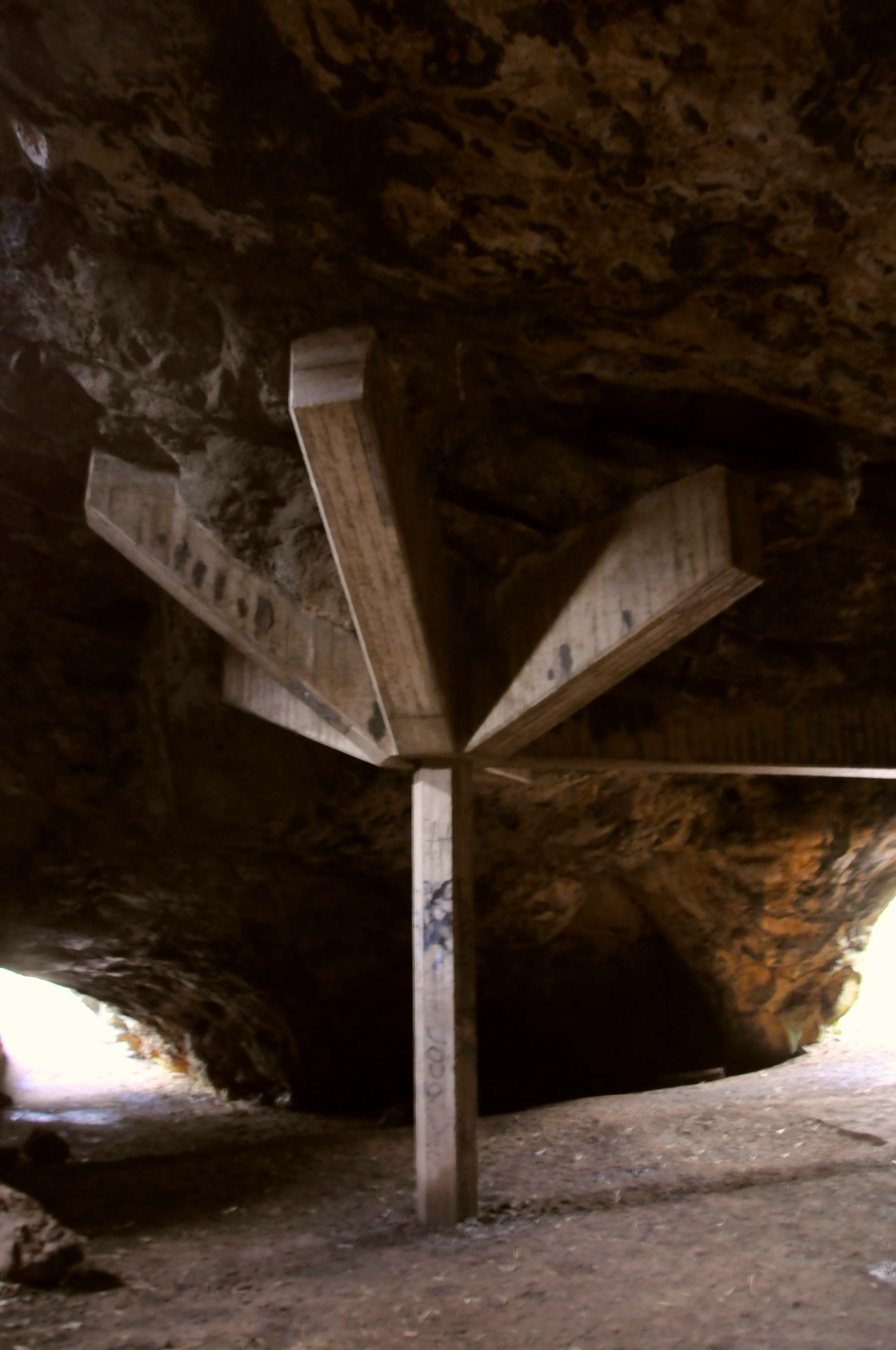 Una gruesa viga de madera de múltiples brazos que sostiene el techo en la entrada de una cueva kárstica, con luz natural entrando por los lados.