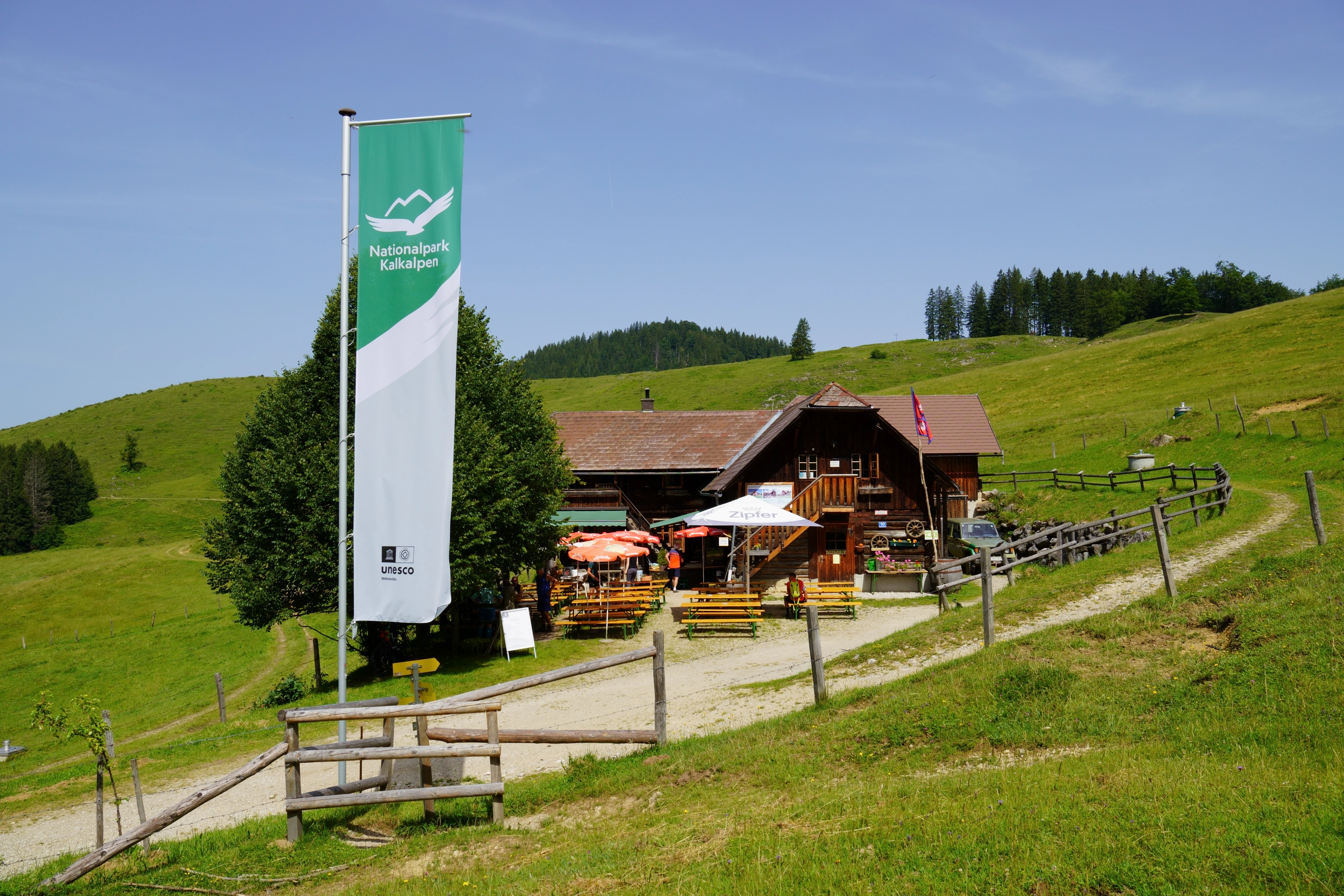 A wooden lodge with outdoor seating, a tall green and white Nationalpark Kalkalpen banner, and a grassy landscape with hills in the background