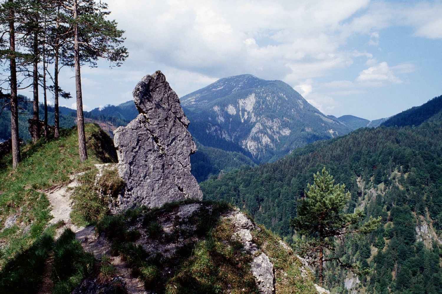 Mountain landscape with rocky outcrop, forested hills, and distant peak under partly cloudy sky