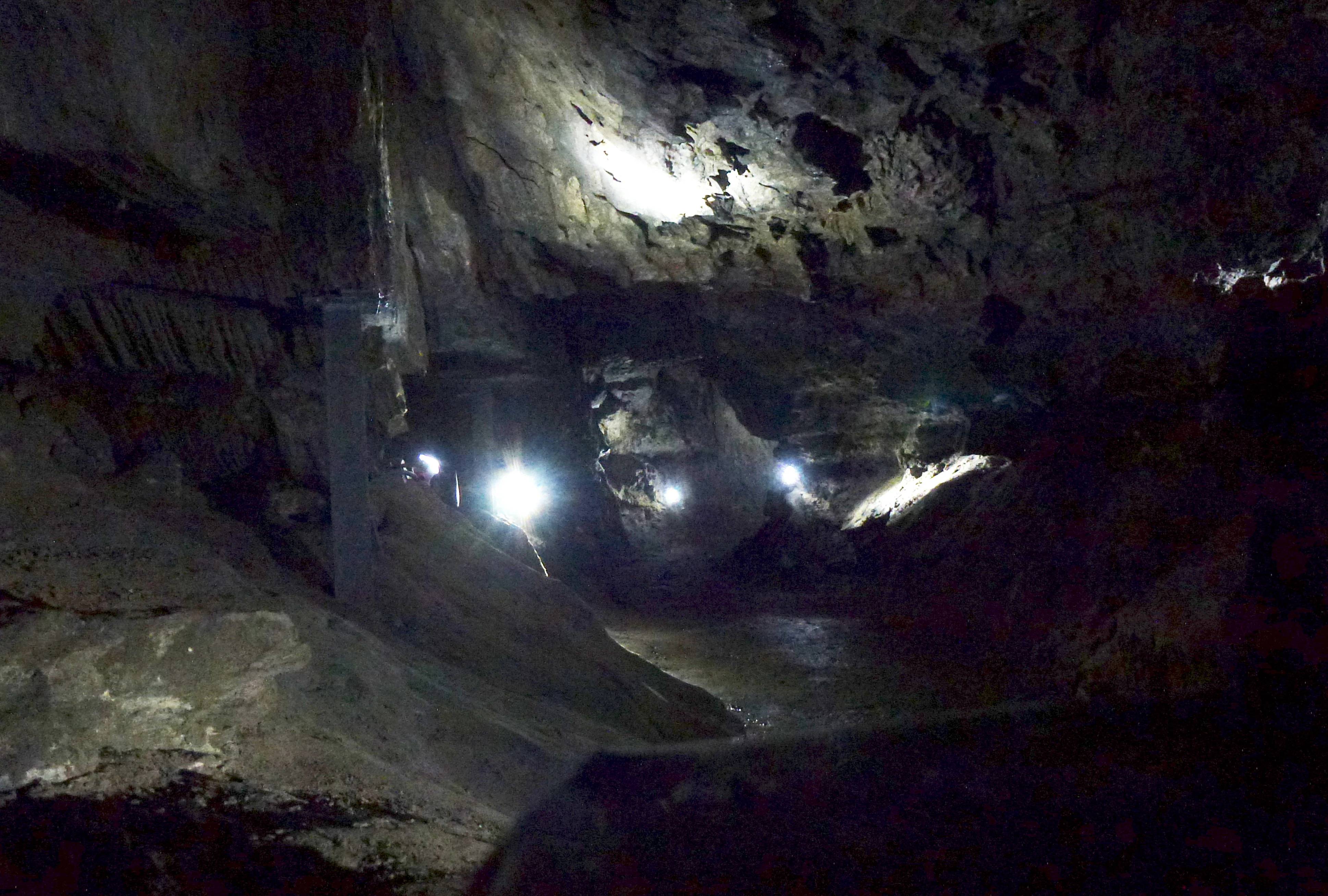 A dimly lit interior view of a natural gypsum cave with rough stone walls, uneven floor, and artificial light sources creating bright spots in the darkness.
