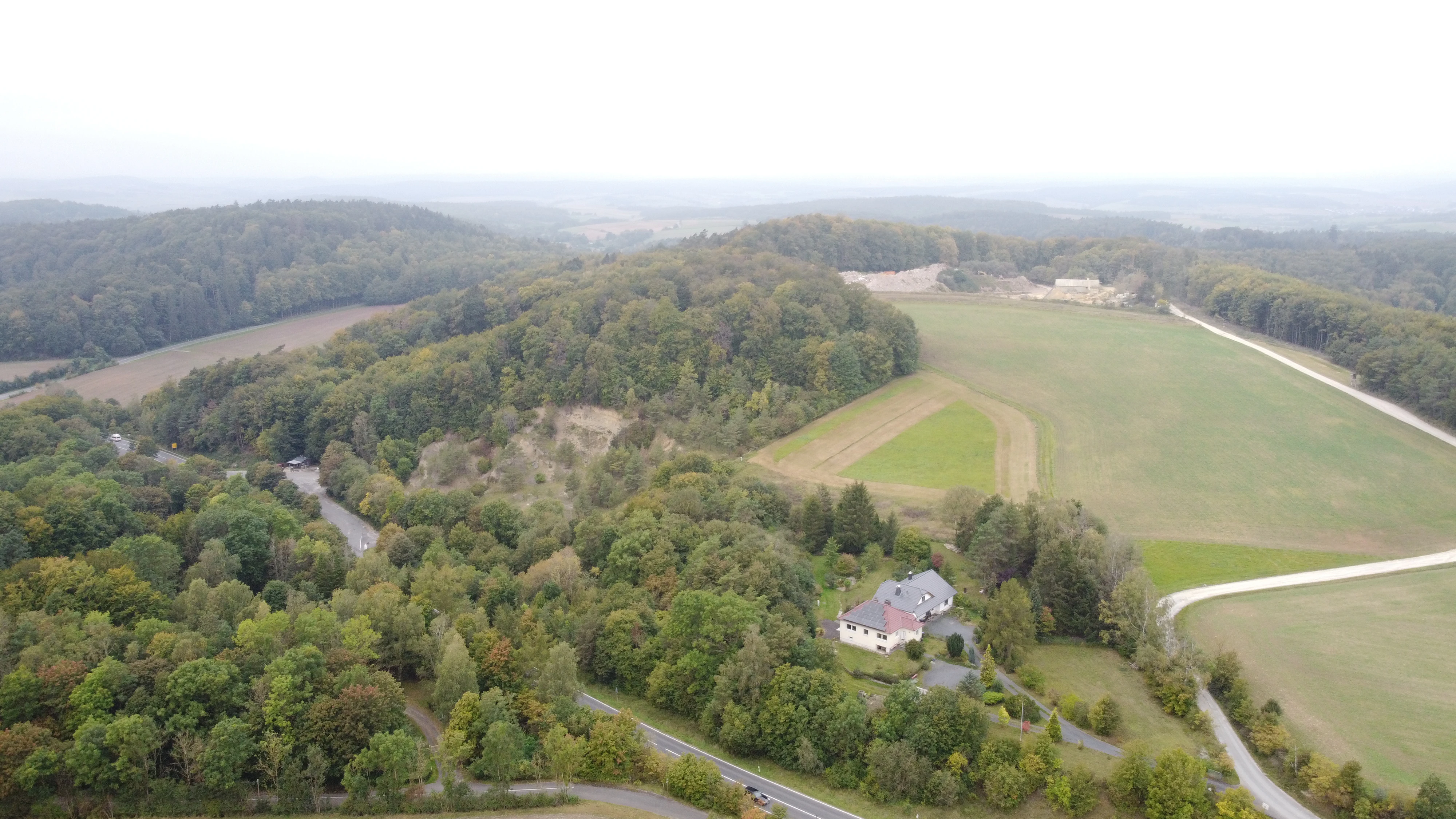 An aerial shot showing forested hills, open agricultural fields, a small cluster of buildings, and a road winding through the German countryside.
