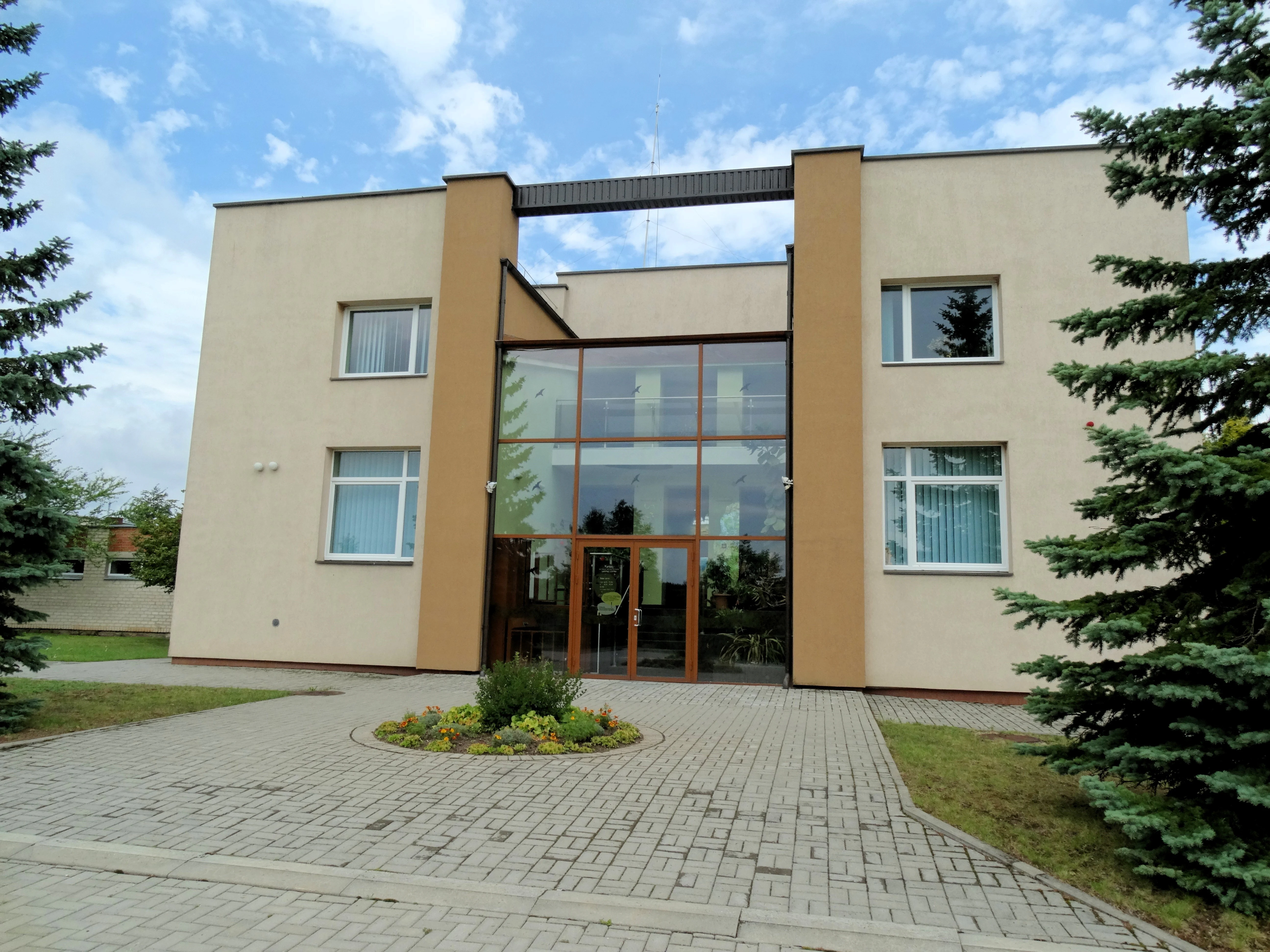 A modern two-story building with a light beige facade, featuring a central glass atrium entrance and a paved courtyard in front.