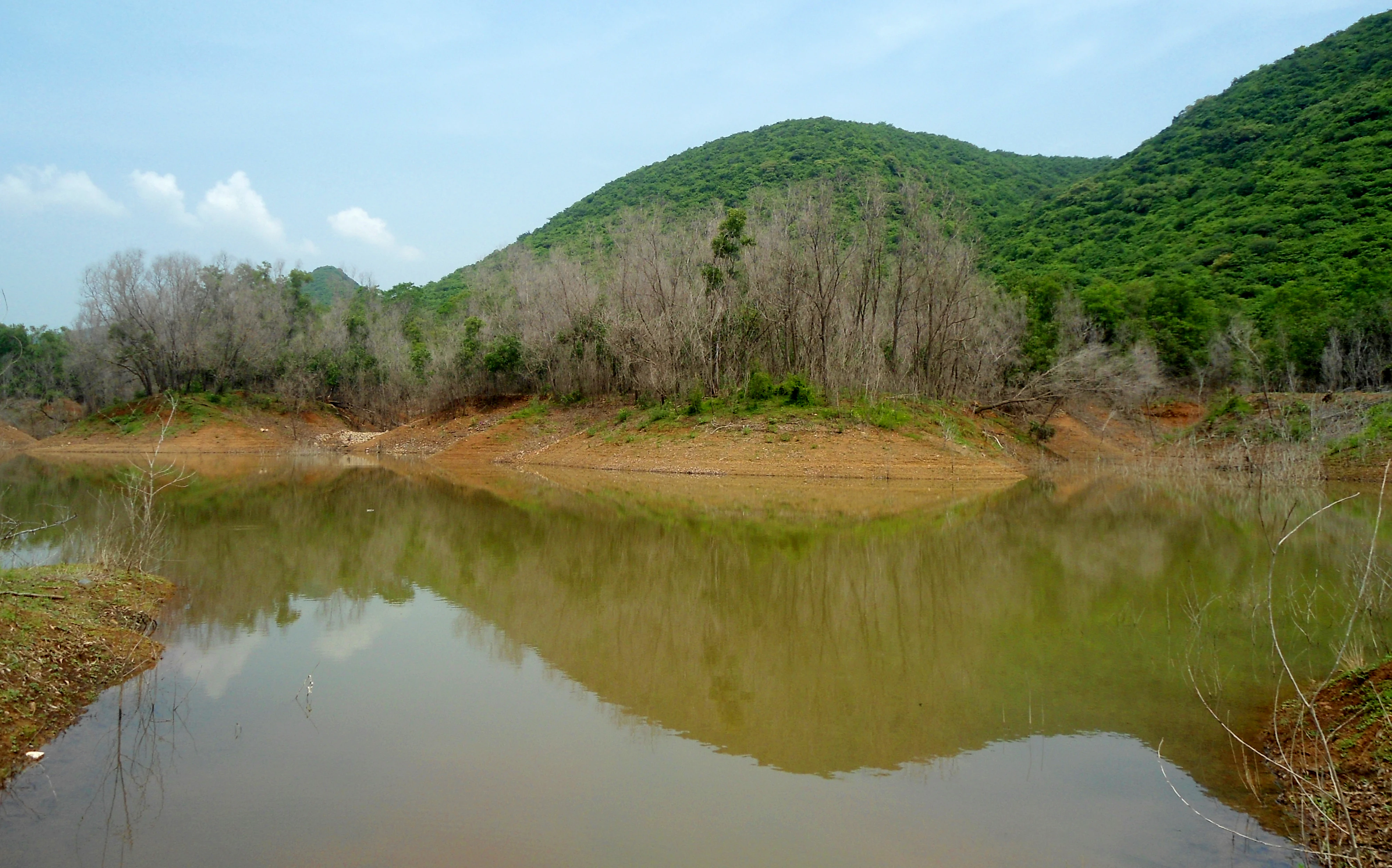A calm, reflective body of water surrounded by green hills and trees that have lost their leaves.