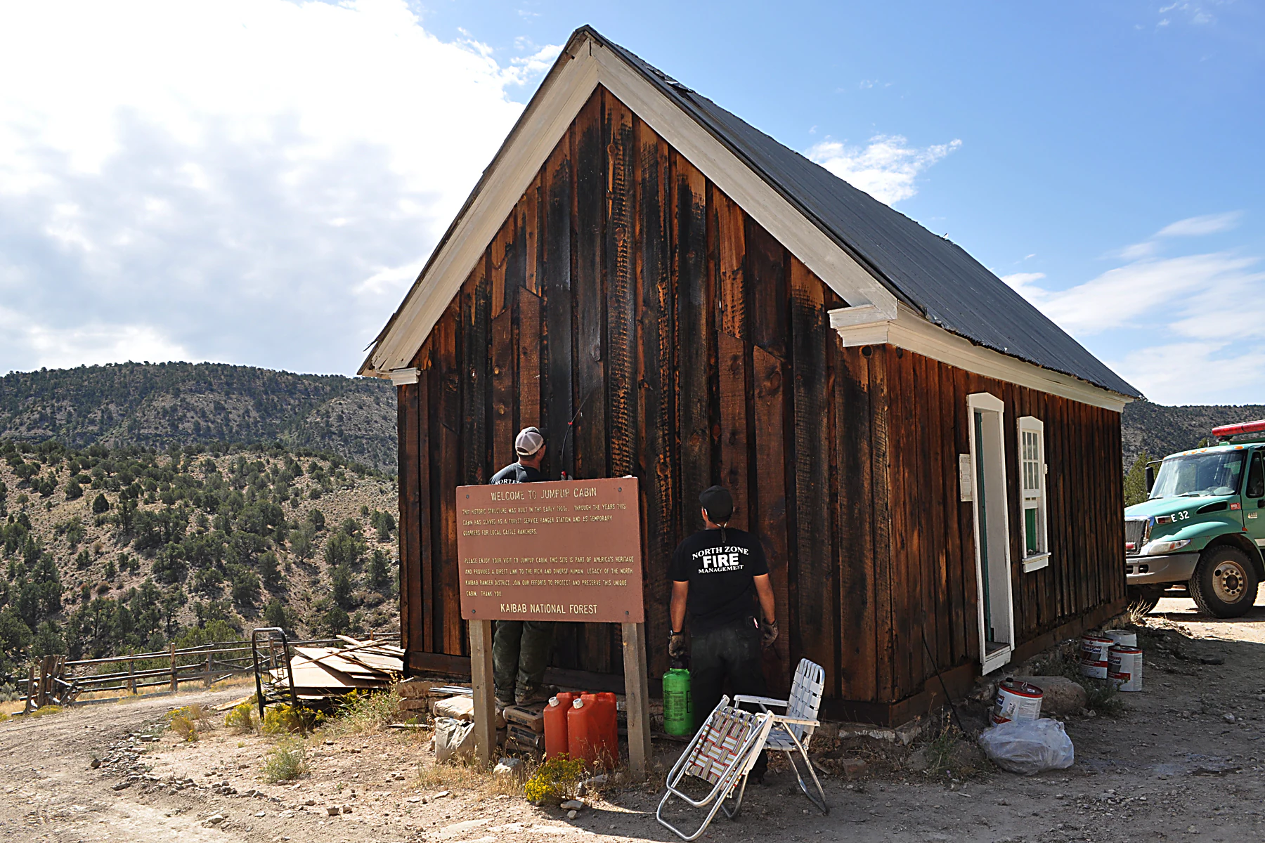 A weathered, dark wooden cabin with a gabled roof stands in a remote landscape, with two workers standing near a wooden historical sign in front of the structure, and a truck parked nearby.