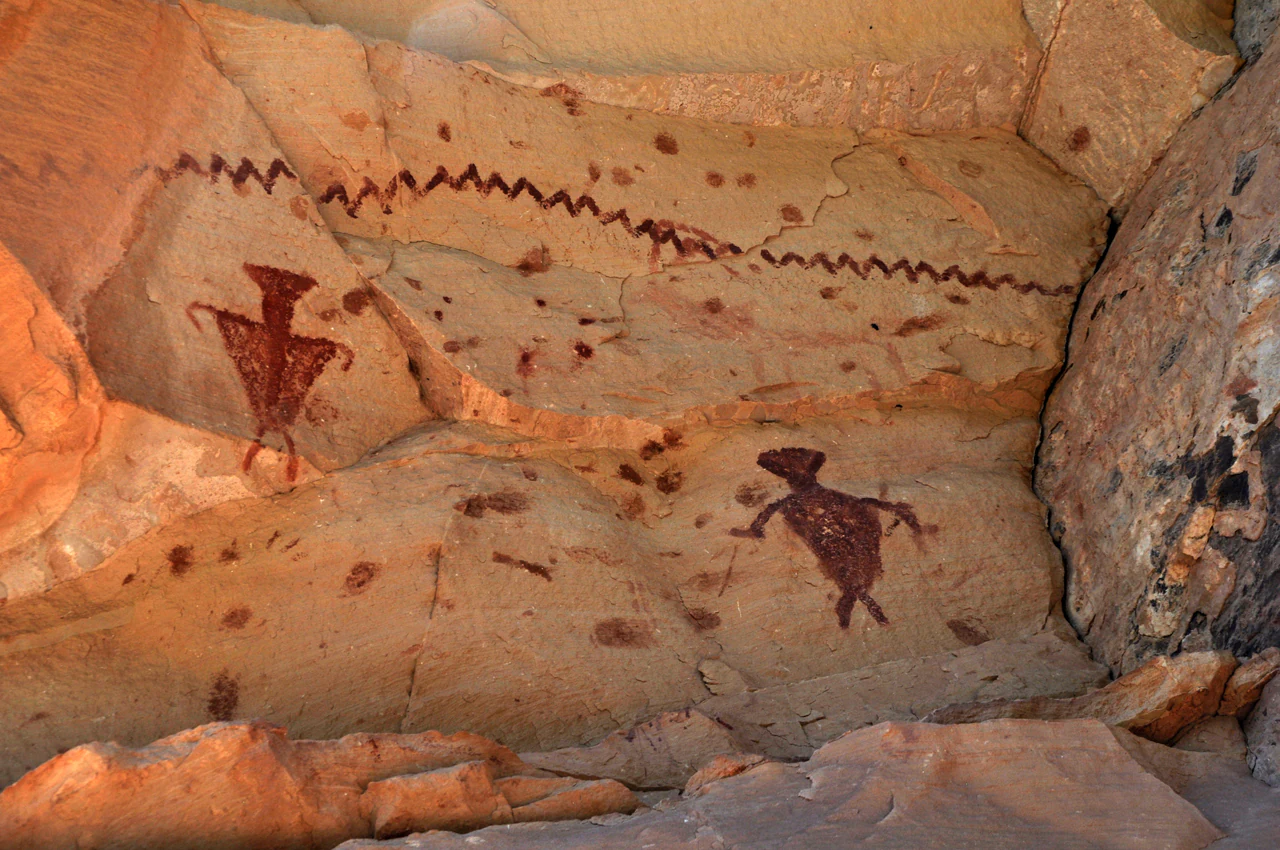 Ancient Native American rock art pictographs of anthropomorphic figures and a zigzag line painted in red pigment on a textured tan sandstone canyon wall.