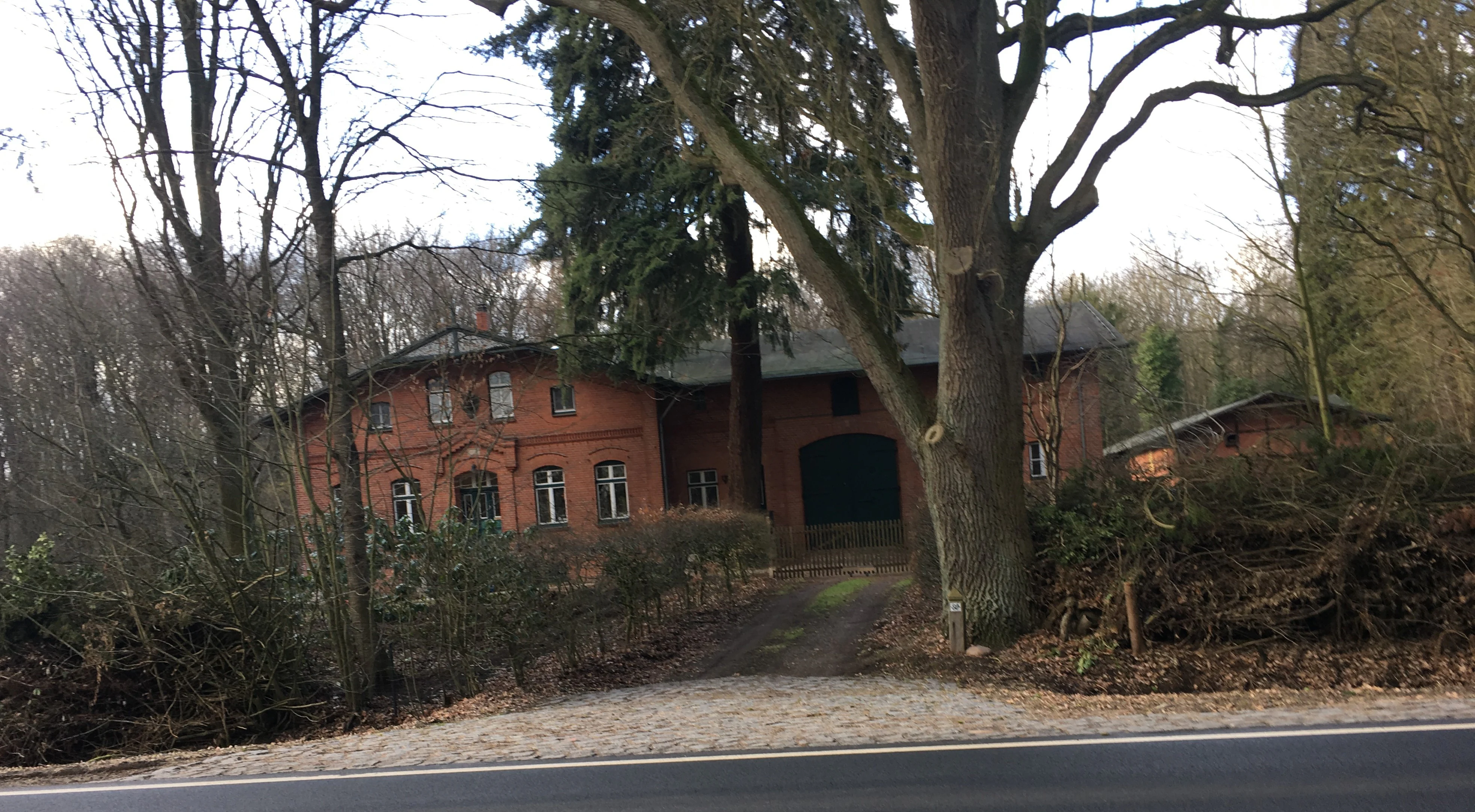 A red brick forestry building viewed from a roadside, framed by large deciduous trees and dense woodland.