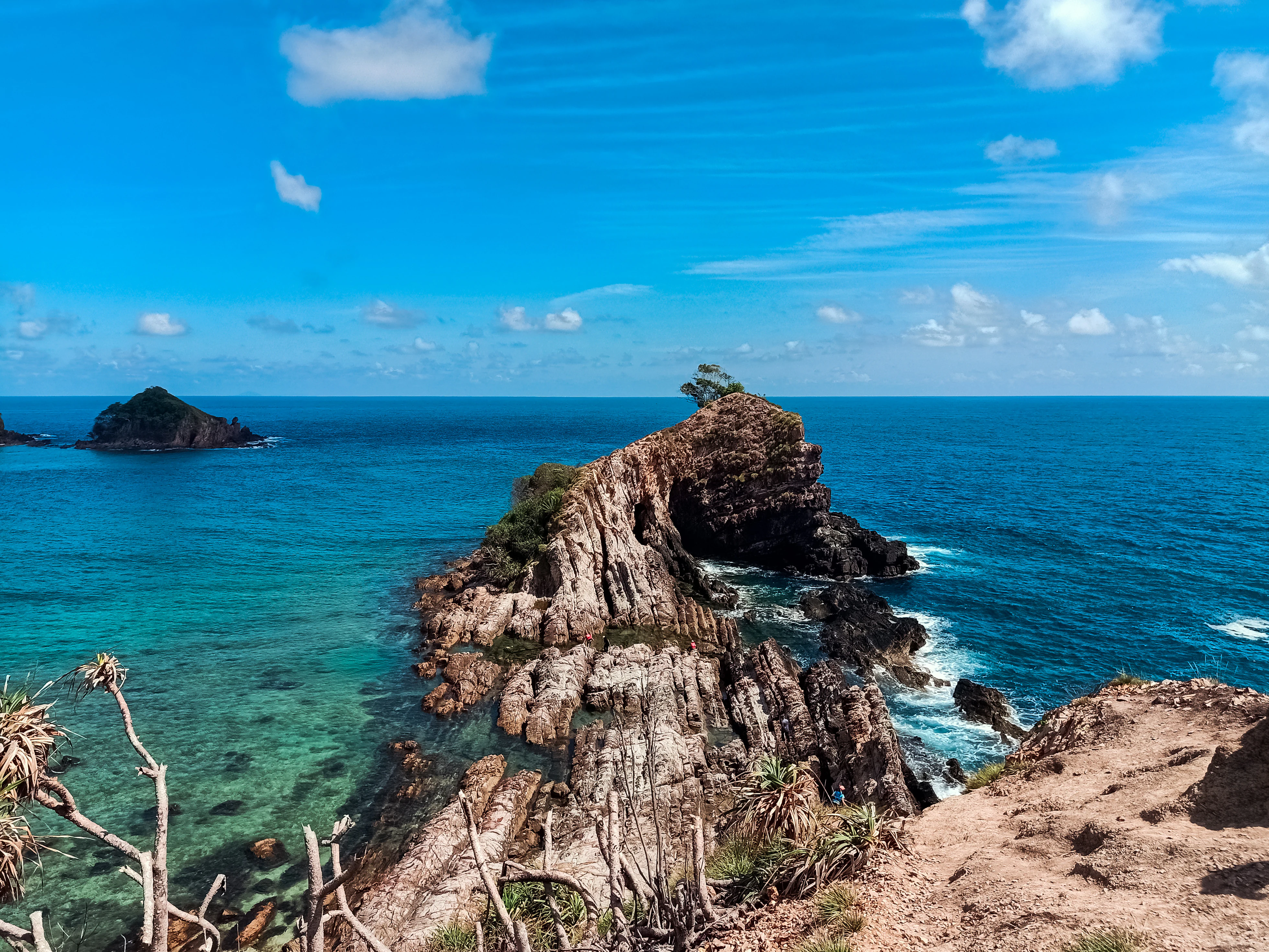 A high-angle view looking over a rugged, rocky ridge protruding into clear, turquoise sea water under a bright blue sky with light clouds.