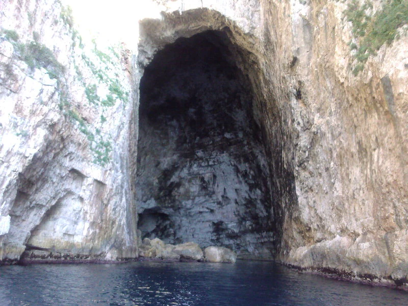 A large, dark cave entrance set into tall, sheer limestone cliffs rising directly out of deep blue sea water.