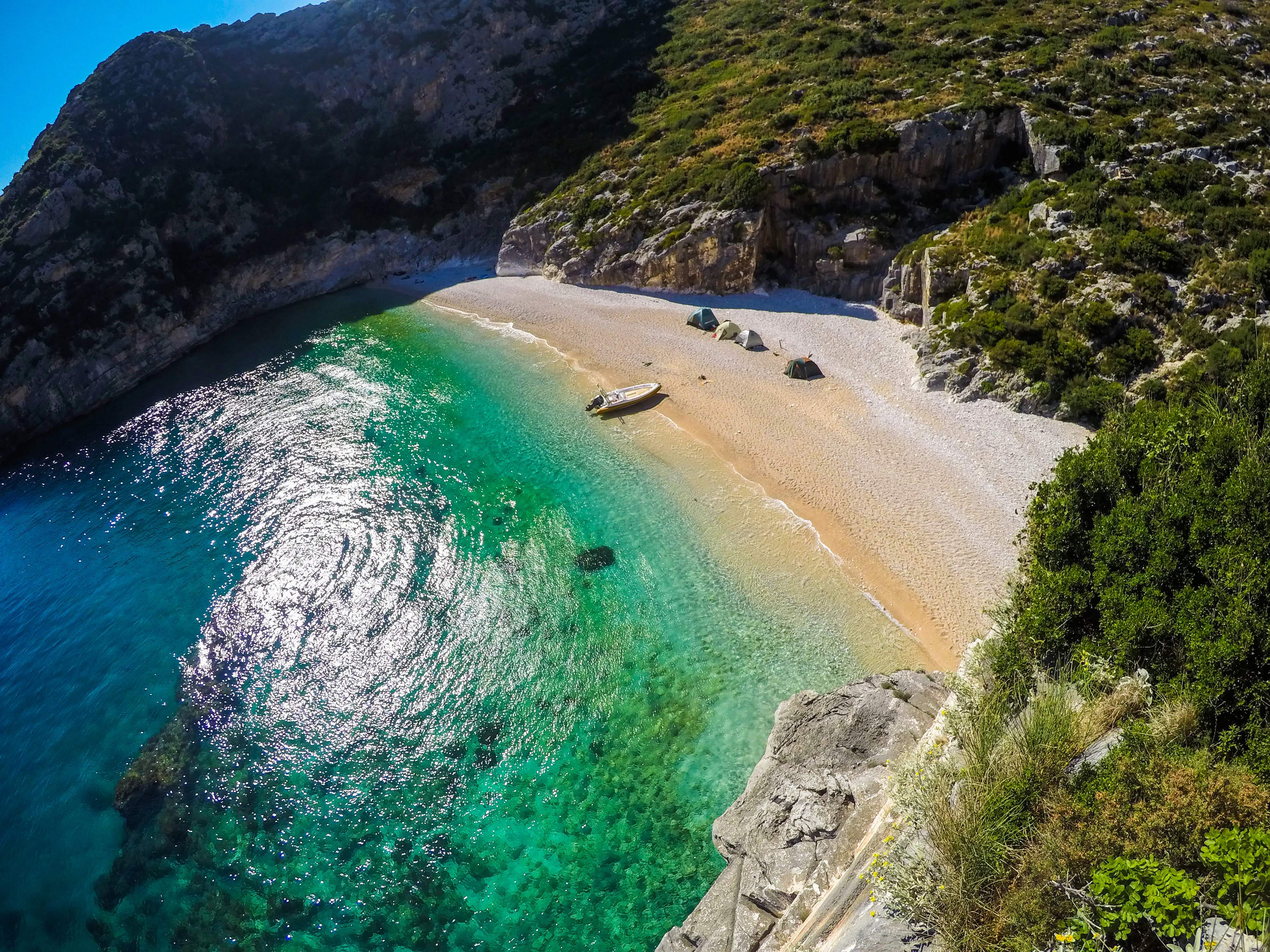 An aerial view of a secluded cove with turquoise water, a pebble beach, and surrounding rocky cliffs covered in green shrubs.