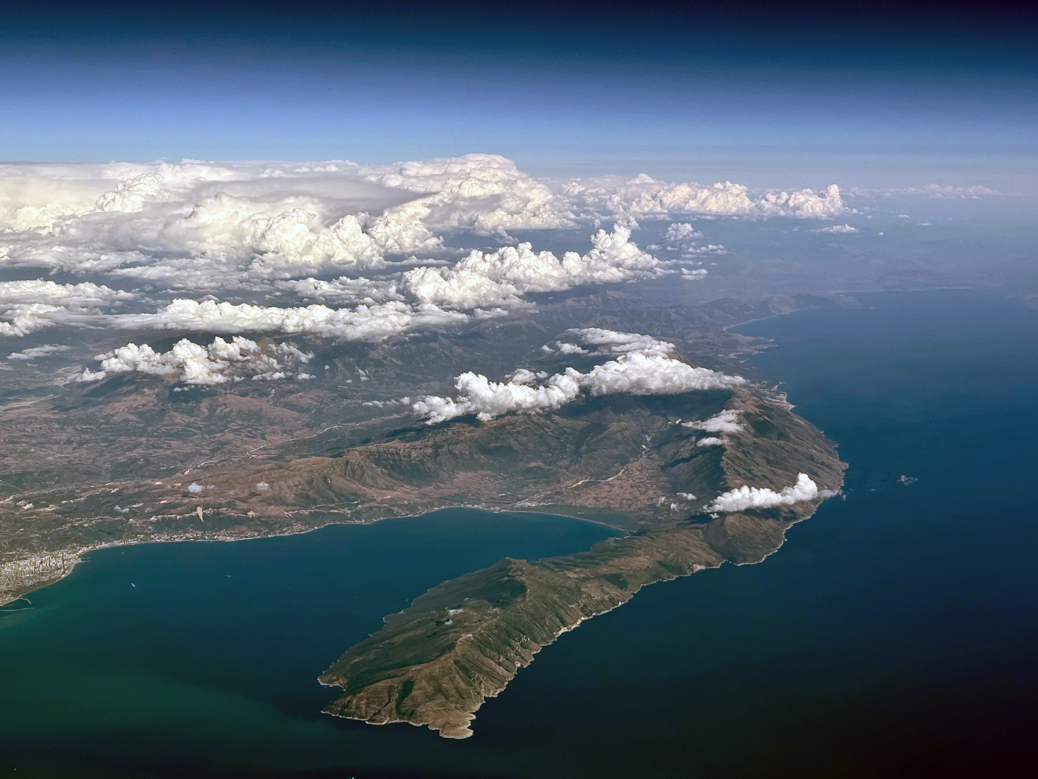 High-altitude aerial view of a large, rugged peninsula extending into deep blue sea, with scattered white clouds over land and mountains.