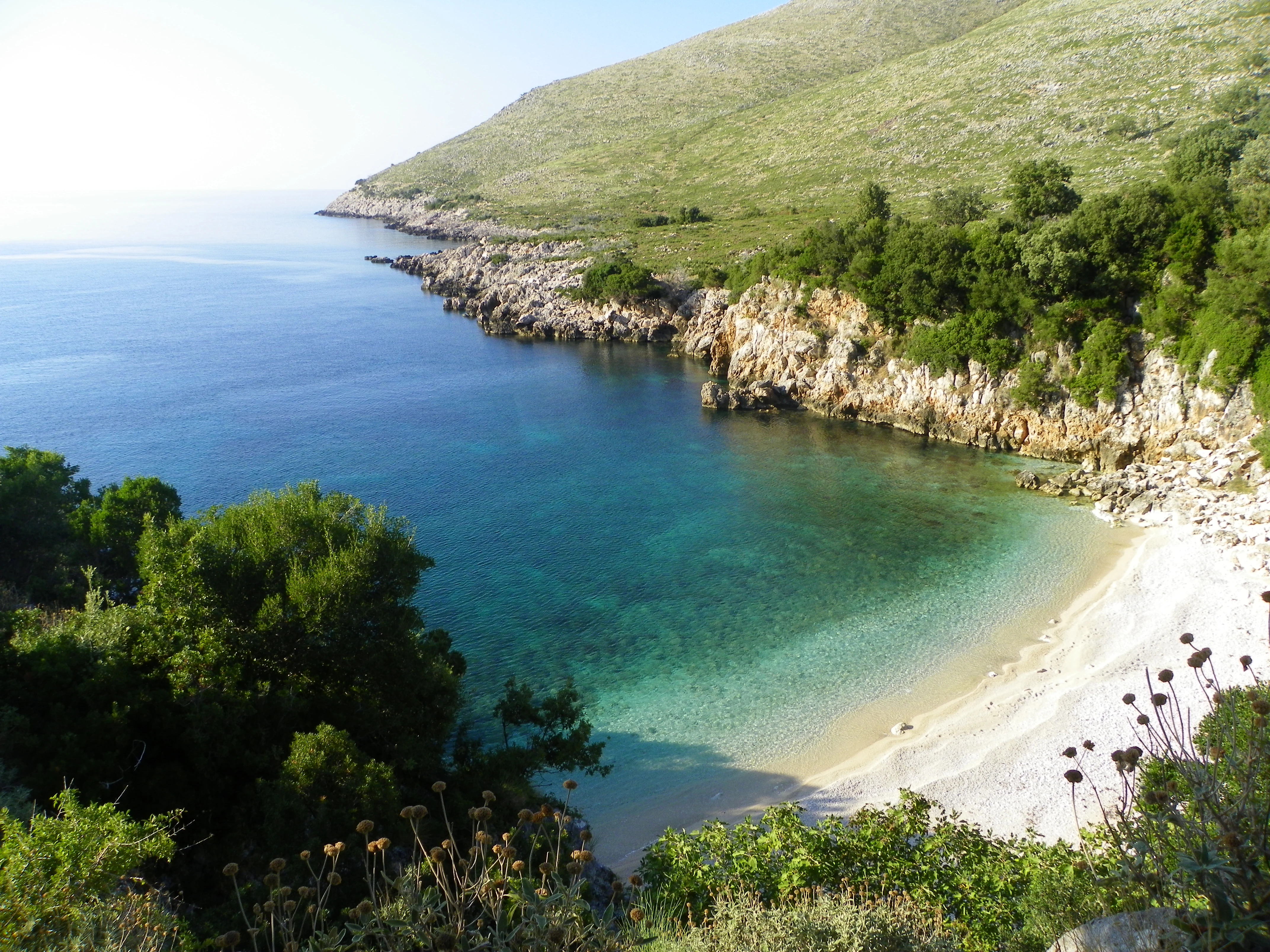 A high-angle view of a secluded cove with a turquoise beach bordered by rocky cliffs and green Mediterranean shrubs.