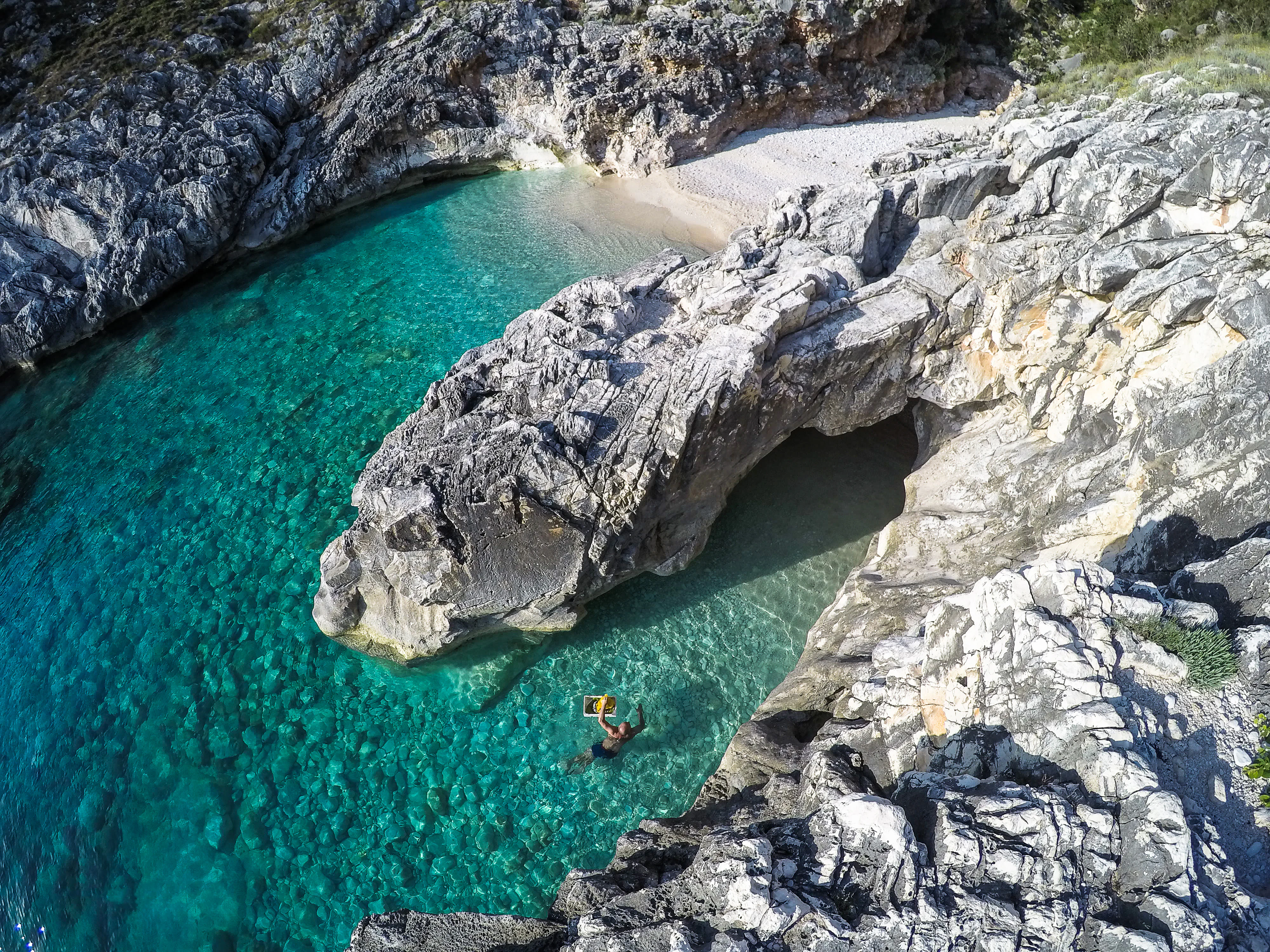 A high-angle view looking down into a small, rocky cove with clear, turquoise water, where a person is swimming; rocky cliffs frame the inlet.