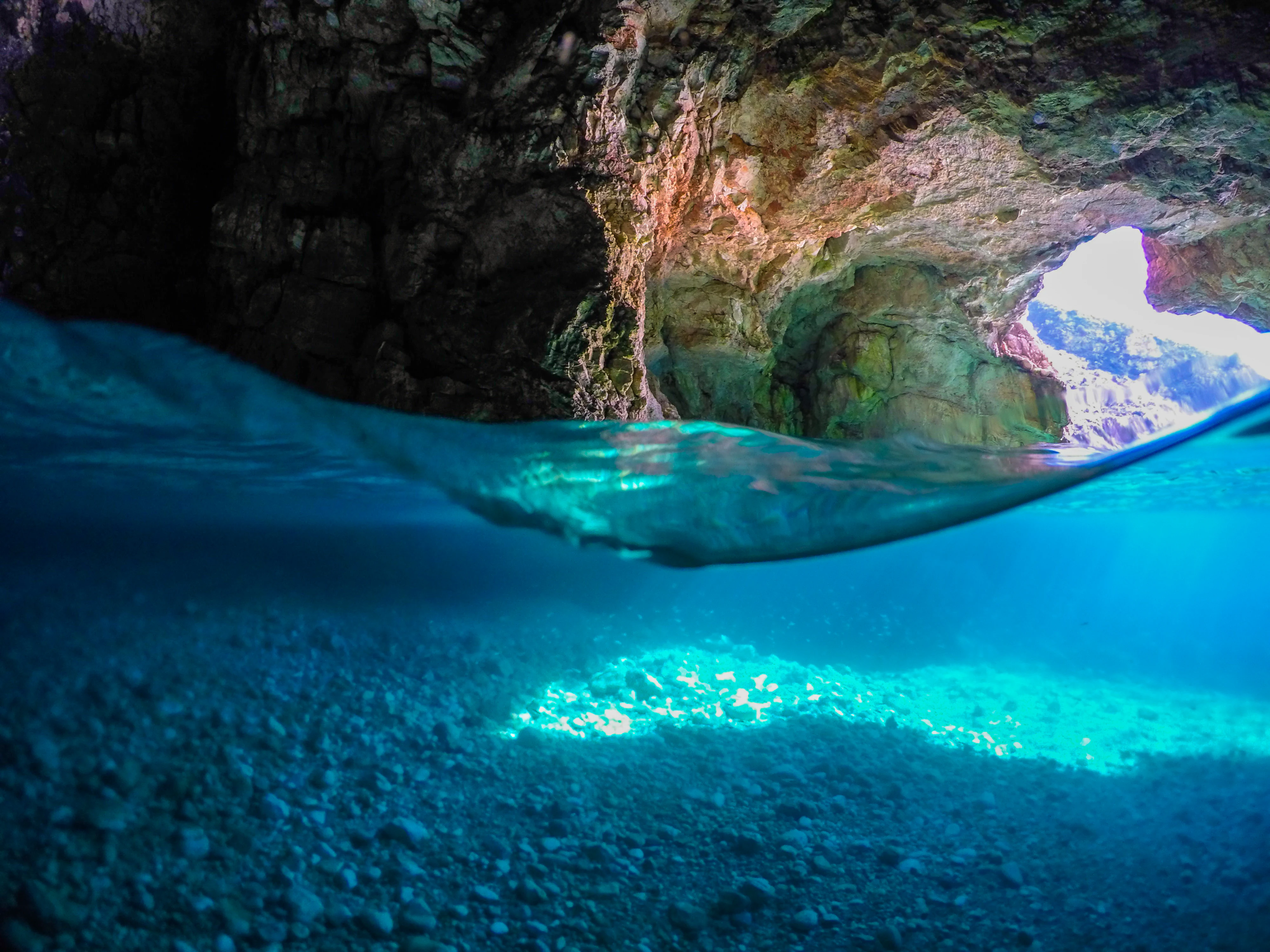 A split-level underwater view of a sea cave. Above the water line, rugged rock formations are visible. Below the water line, the seafloor consists of light-colored pebbles illuminated by blue light.