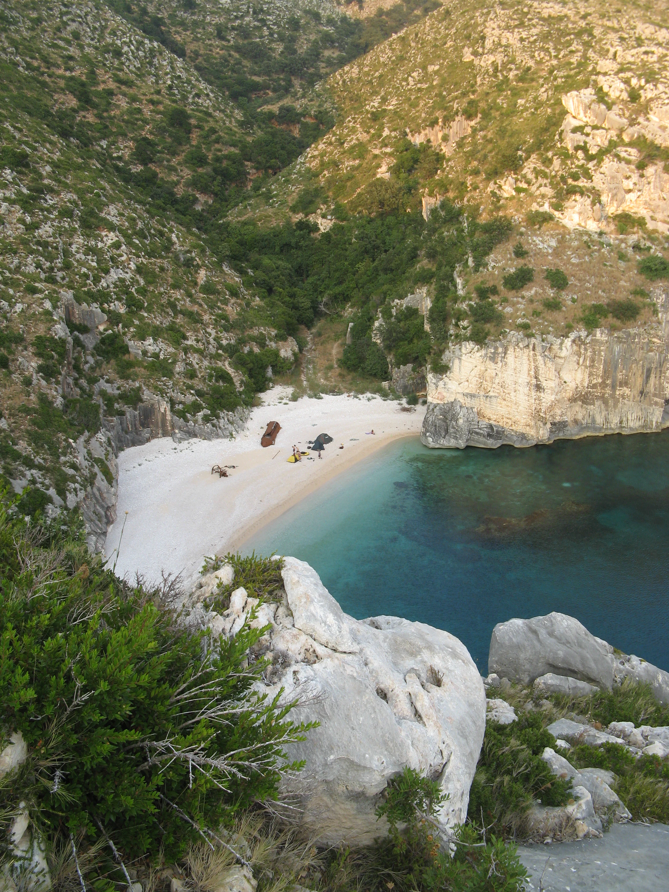 High-angle view of a secluded white pebble beach at Grama Bay, nestled between steep limestone cliffs and turquoise water.