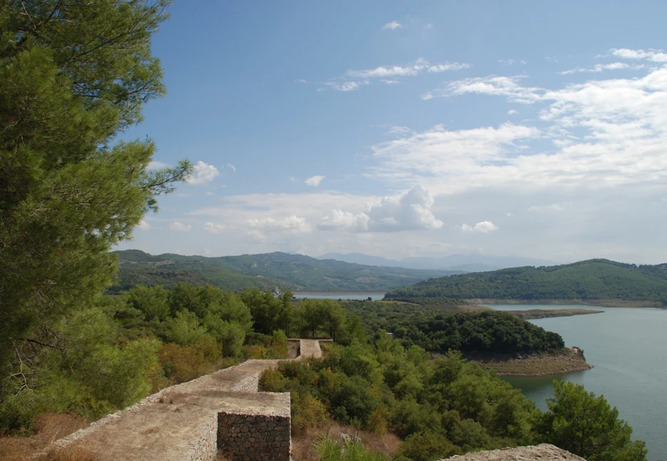 Stone city wall running along a hillside with a reservoir and mountains in the background