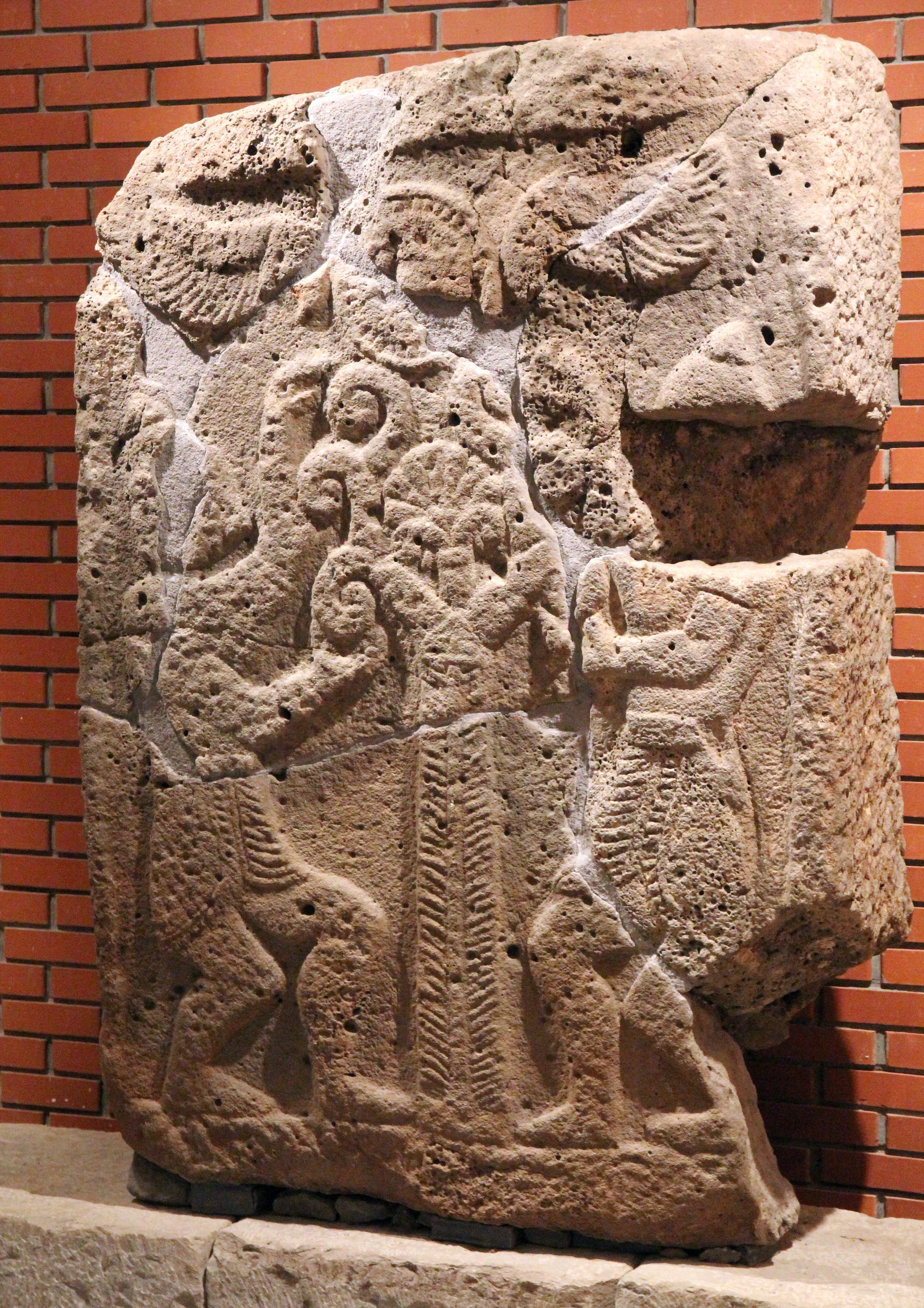Stone relief carving depicting figures dancing around a tree of life with a sun disk, displayed against a brick wall