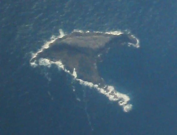 An aerial photograph looking down at a small, dark, rocky island surrounded by dark blue ocean water with white surf breaking against the shoreline.