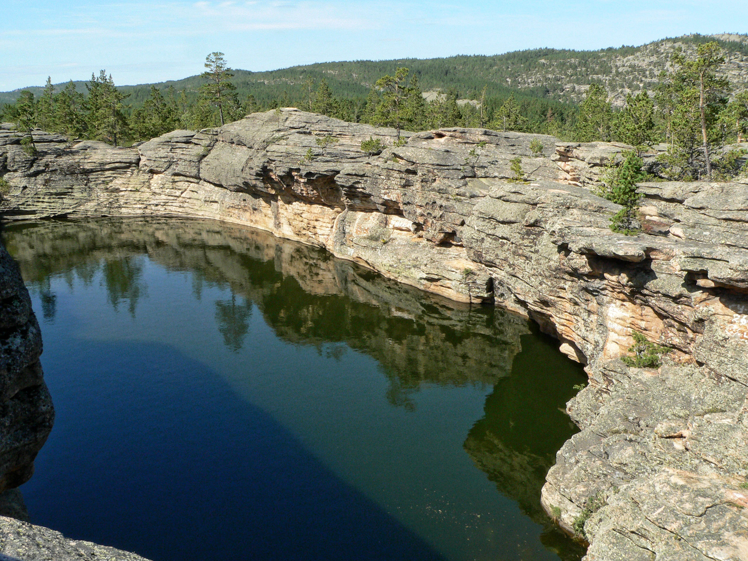 Calm lake reflecting rocky cliffs and forested hills under a clear sky