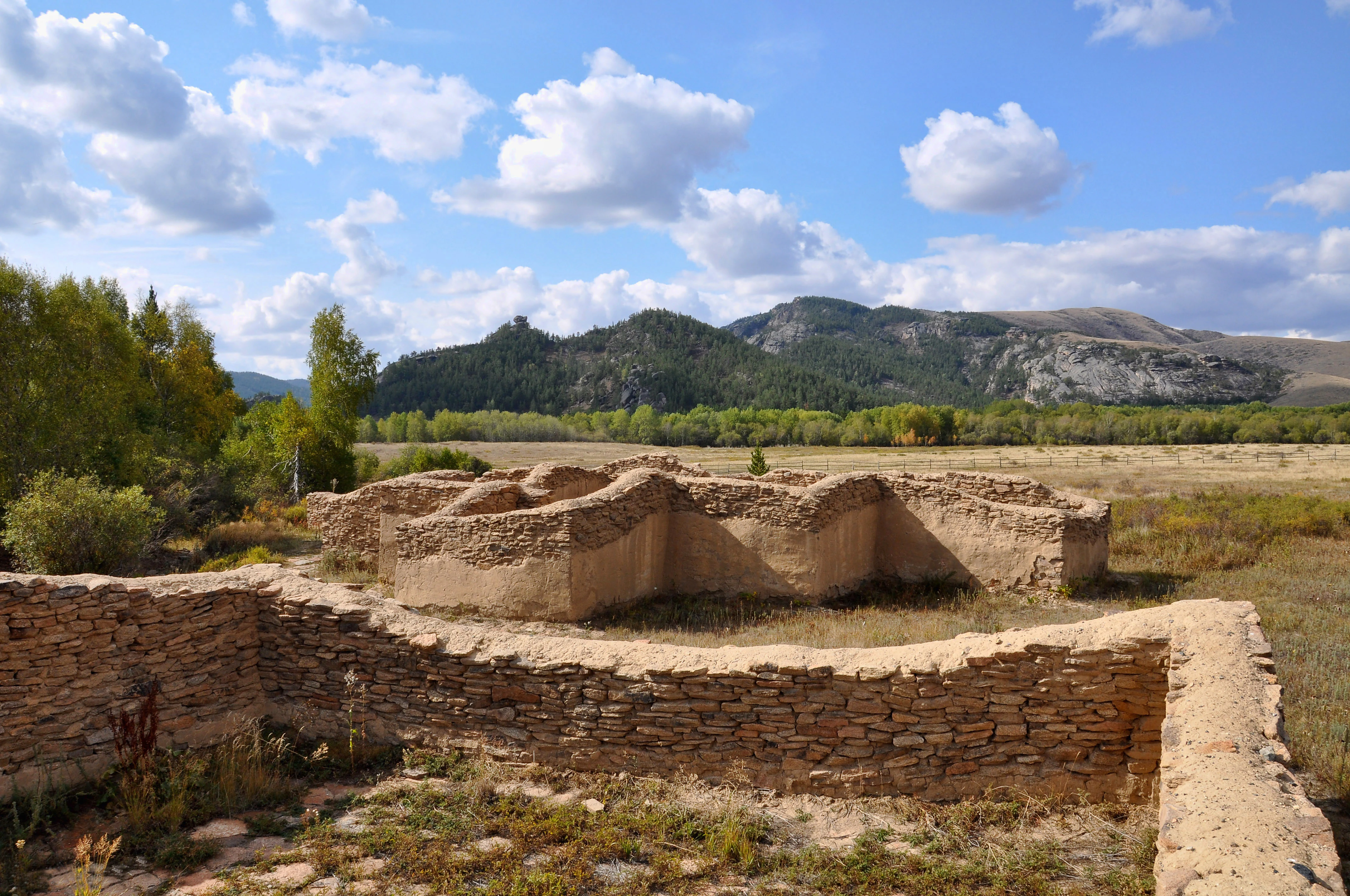 Stone ruins of an ancient palace structure surrounded by grassland and mountains under a partly cloudy sky