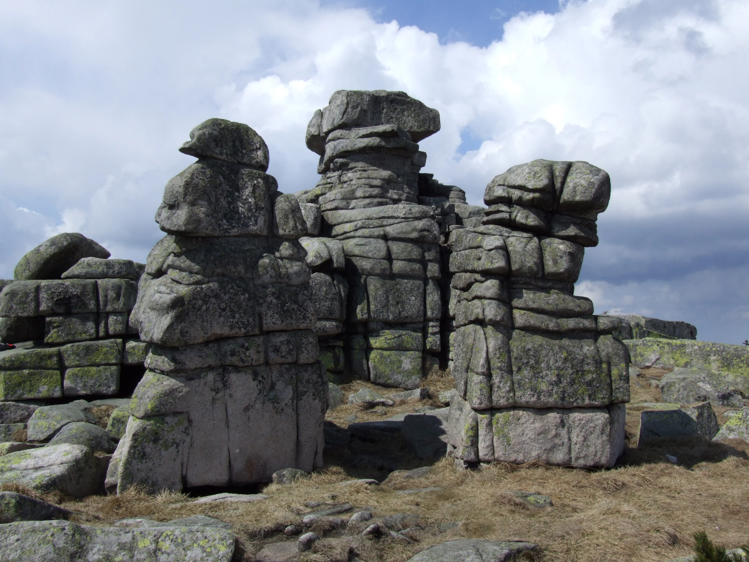 A group of weathered, blocky granite rock formations rising from a dry, grassy mountain landscape under a partly cloudy sky.