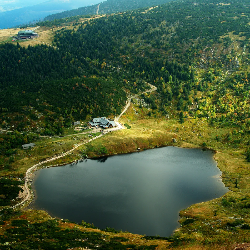 Aerial view of a mountain lake surrounded by forests and hills with a mountain hut and dirt paths nearby