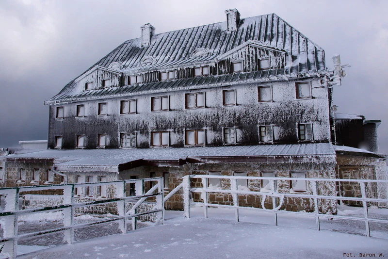 Snow-covered stone mountain refuge building with multiple windows and metal railing, set against a cloudy sky