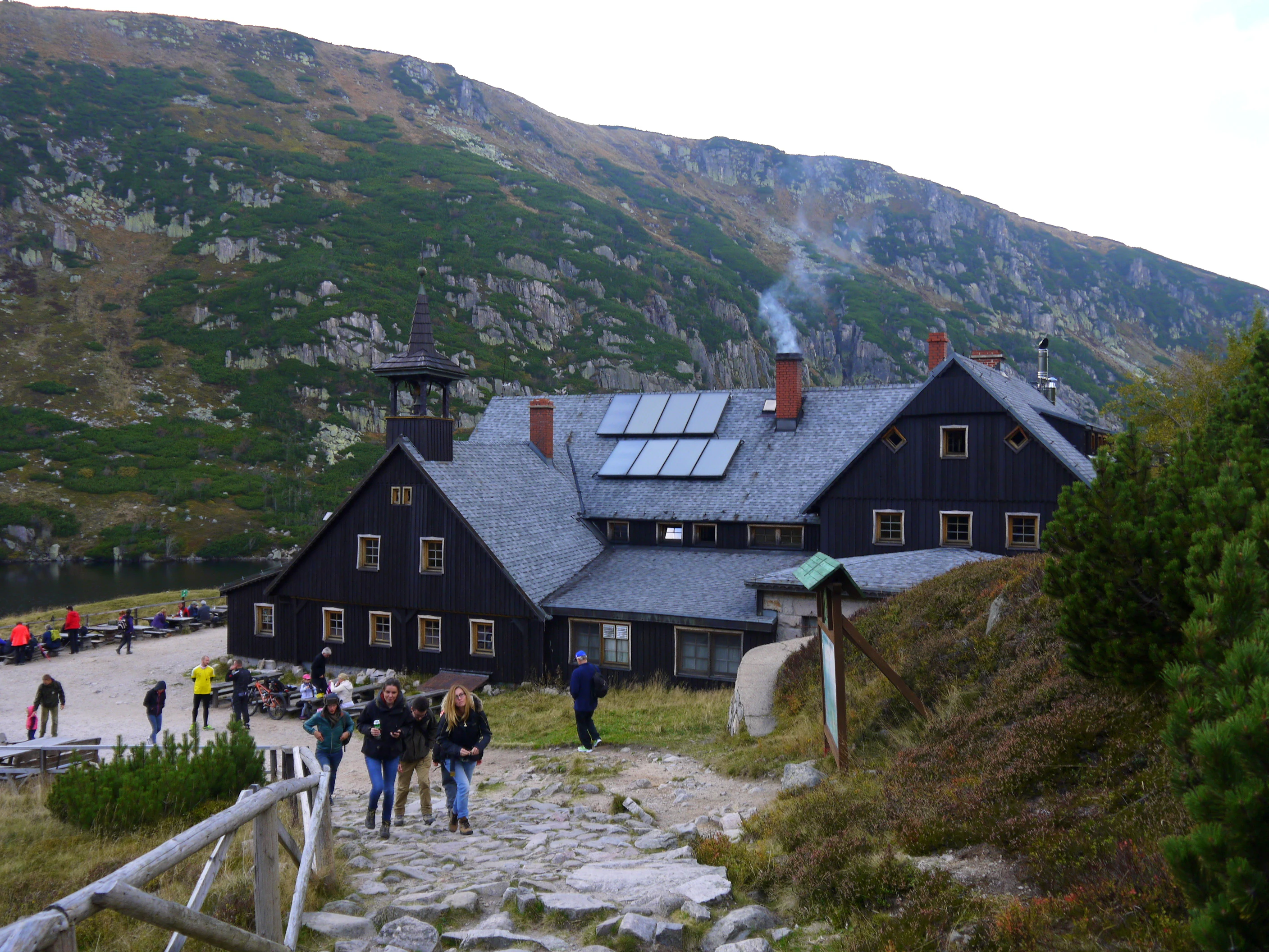 Dark wooden mountain lodge building with solar panels on roof, stone path leading to entrance, people walking on path, mountains and body of water in background