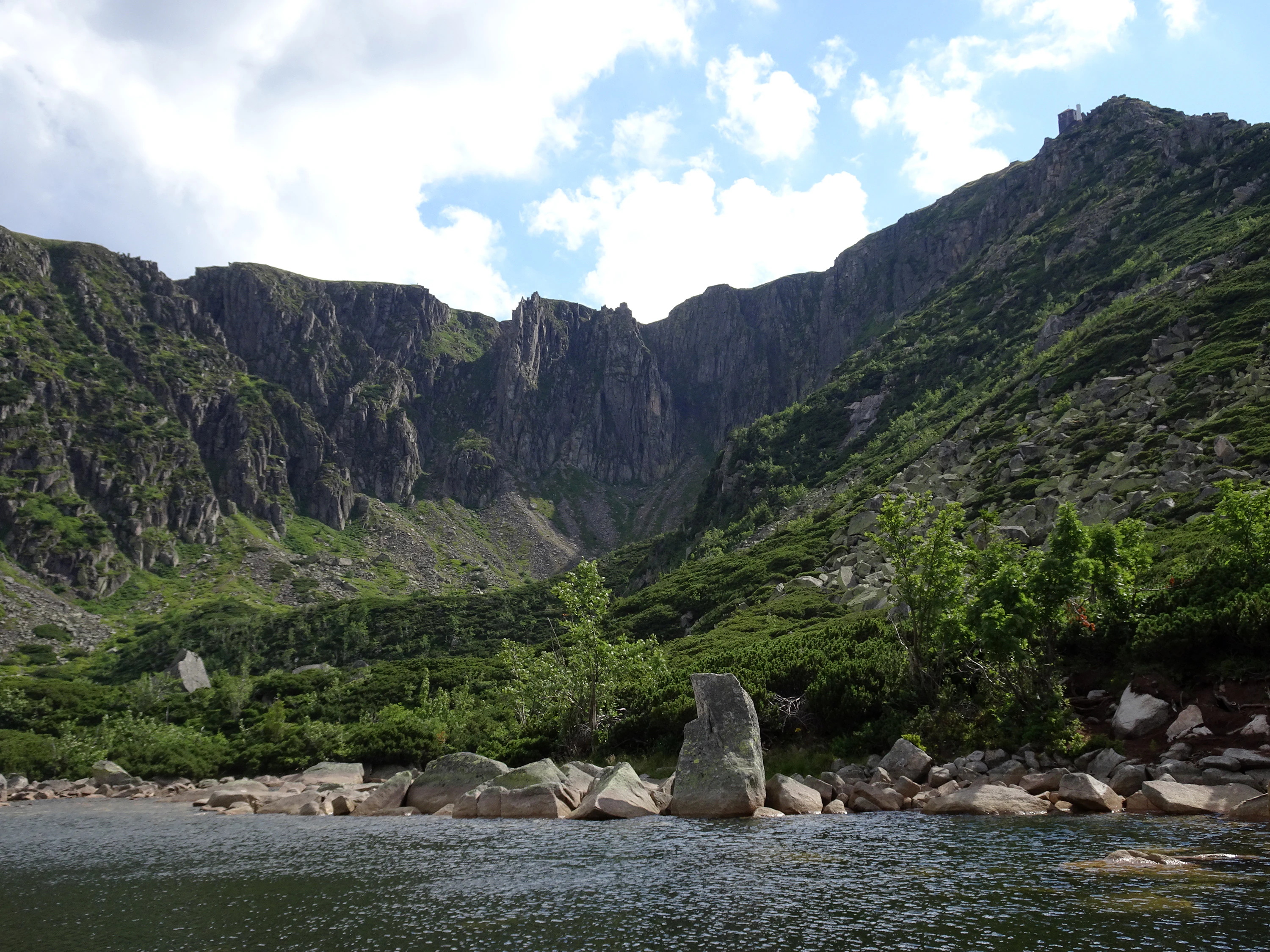 Mountain lake with rocky shore, tall rock formation, green forested slopes, and rocky peaks under partly cloudy sky
